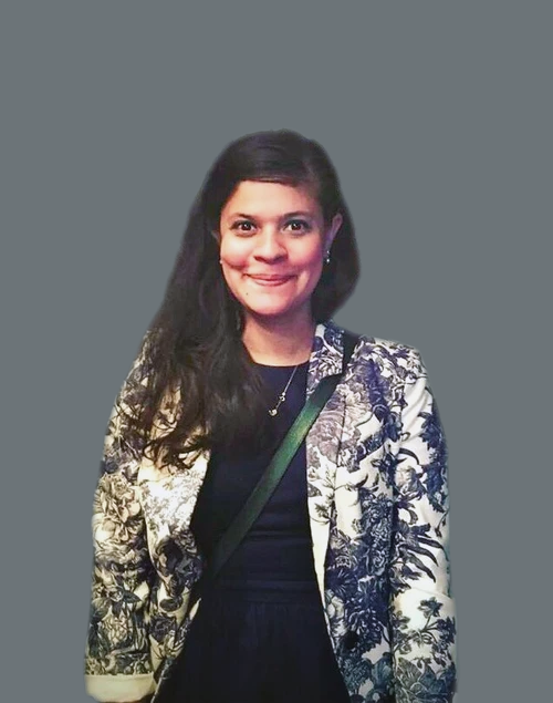 Portrait of Amanda Marikar with dark hair smiling, wearing a black top and a patterned blazer with black and white floral designs, standing against a plain gray background.