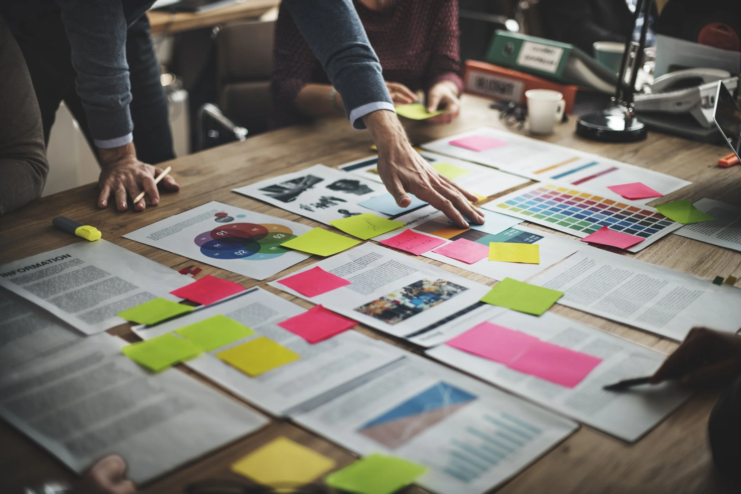 People discussing and analyzing documents, charts, and color palettes on a wooden table during a meeting or project planning session.