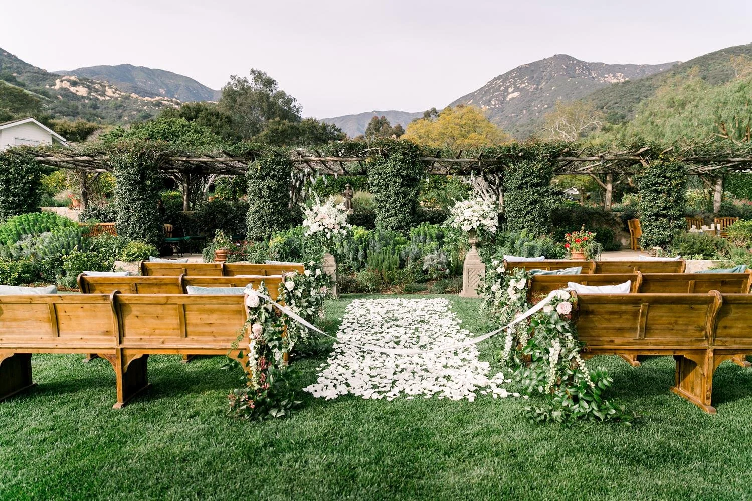 Wedding ceremony in front of arbor with white florals, greenery, and pews at San Ysidro Ranch