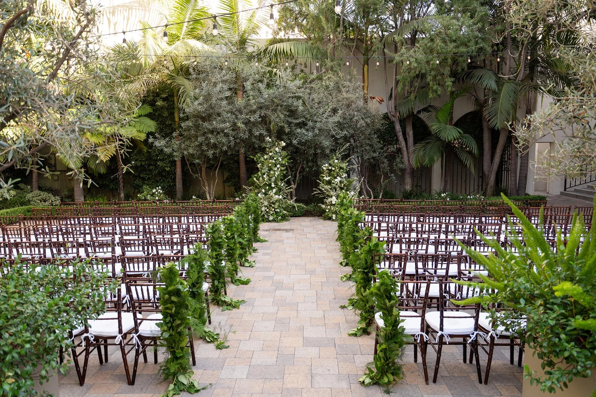 Wide view of a courtyard wedding ceremony with a green garland-lined aisle at Vibiana in Los Angeles