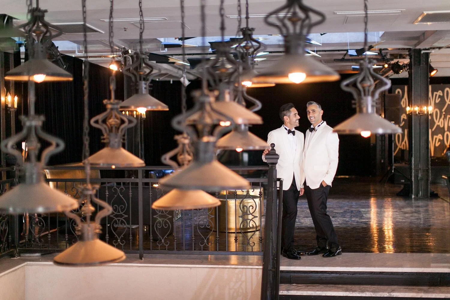 Wedding portrait of grooms standing atop stairs beneath industrial chandeliers at the Alexandria Ballrooms