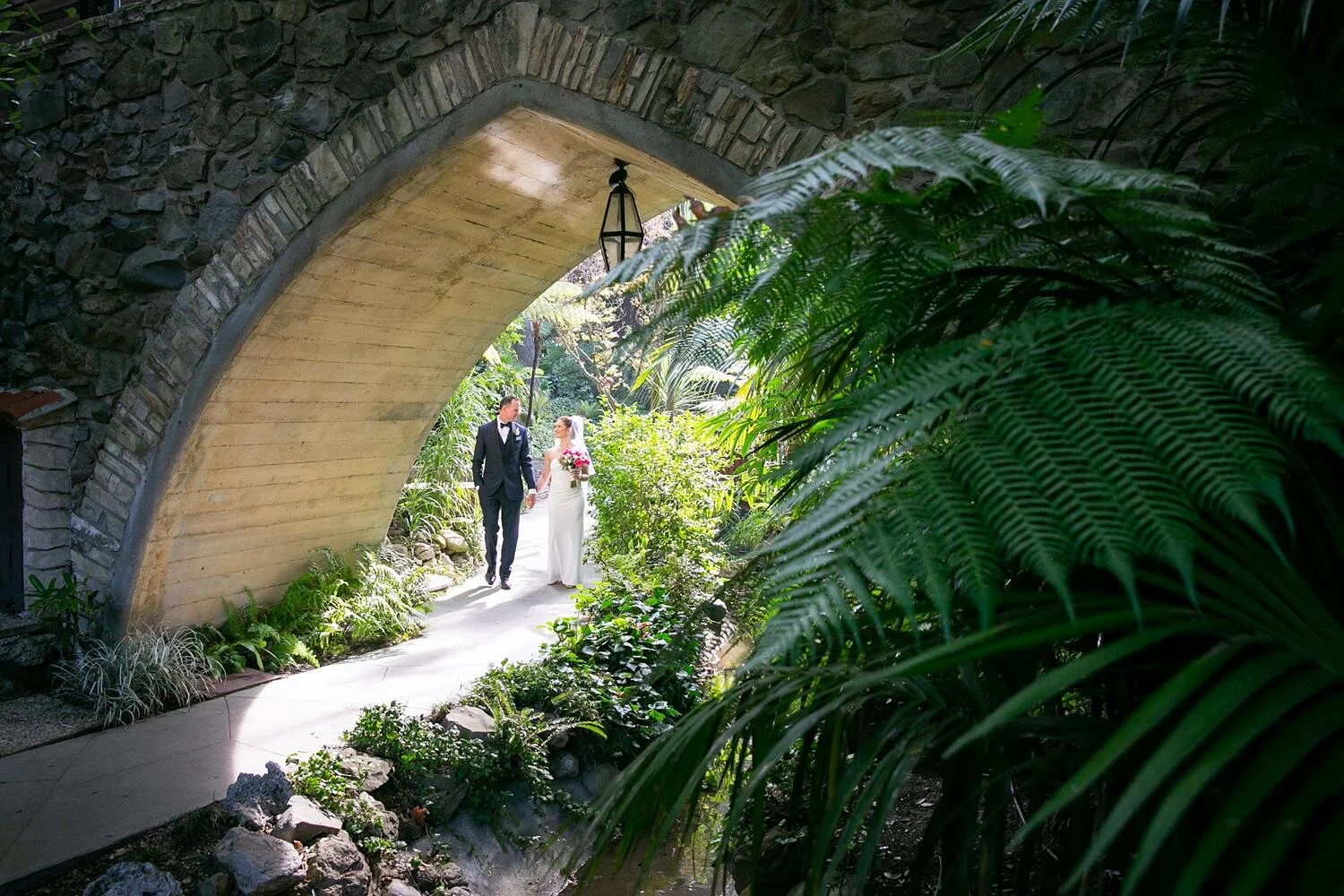 Wedding portraits of groom walking beneath the fern-lined bridge at Hotel Bel Air