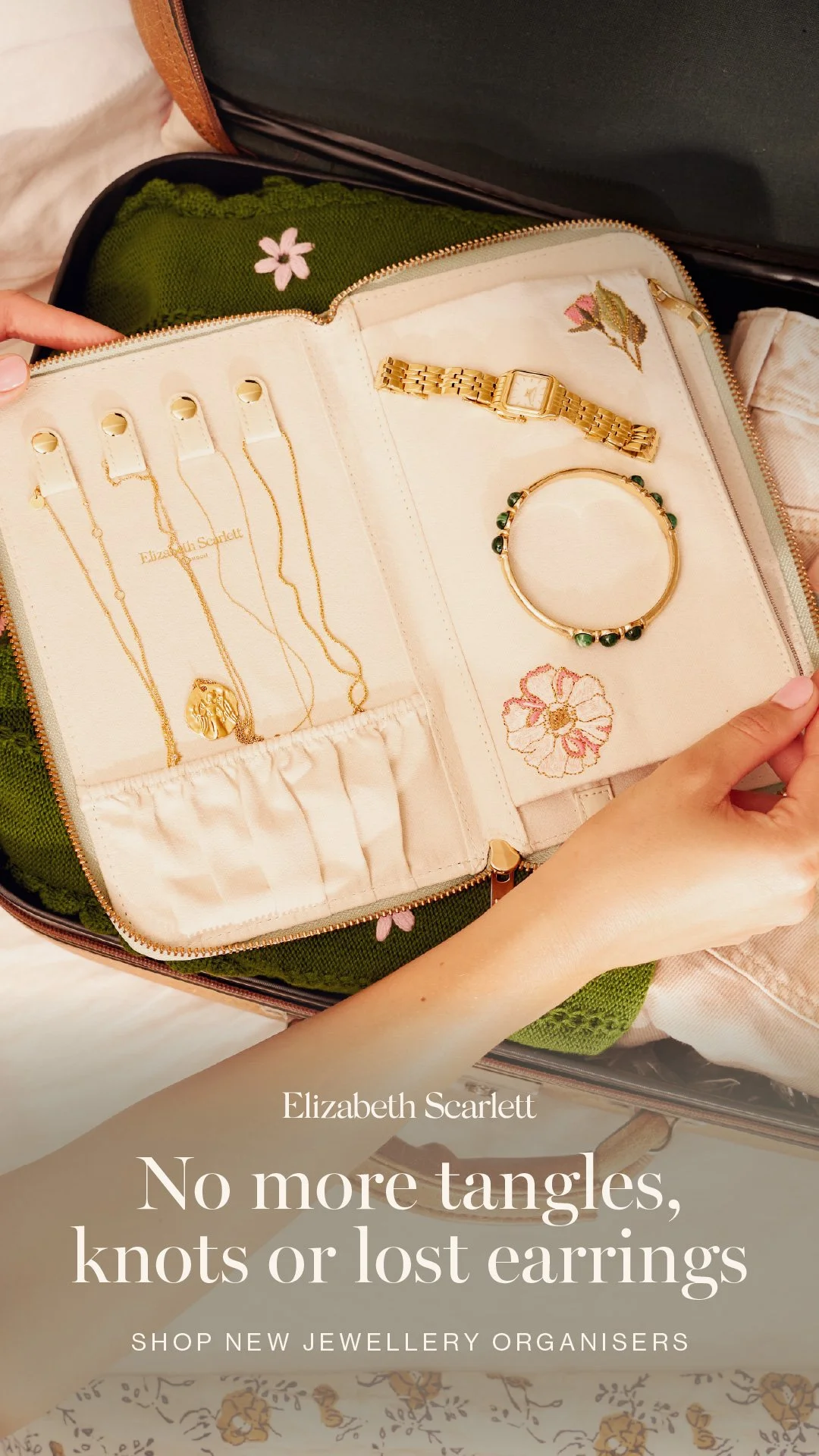 An open jewelry organiser case with gold jewellery including necklaces, a watch, a bracelet, and a ring, with embroidered floral details inside. A person's hand is visible holding the case.