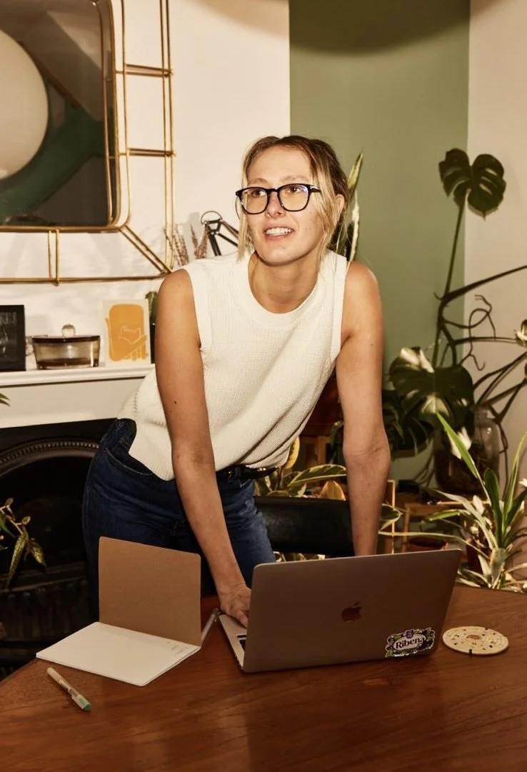 A woman with short hair, glasses, wearing a sleeveless white top, leaning over a wooden table in a cozy indoor setting with plants and decor, with a laptop and notebook on the table.