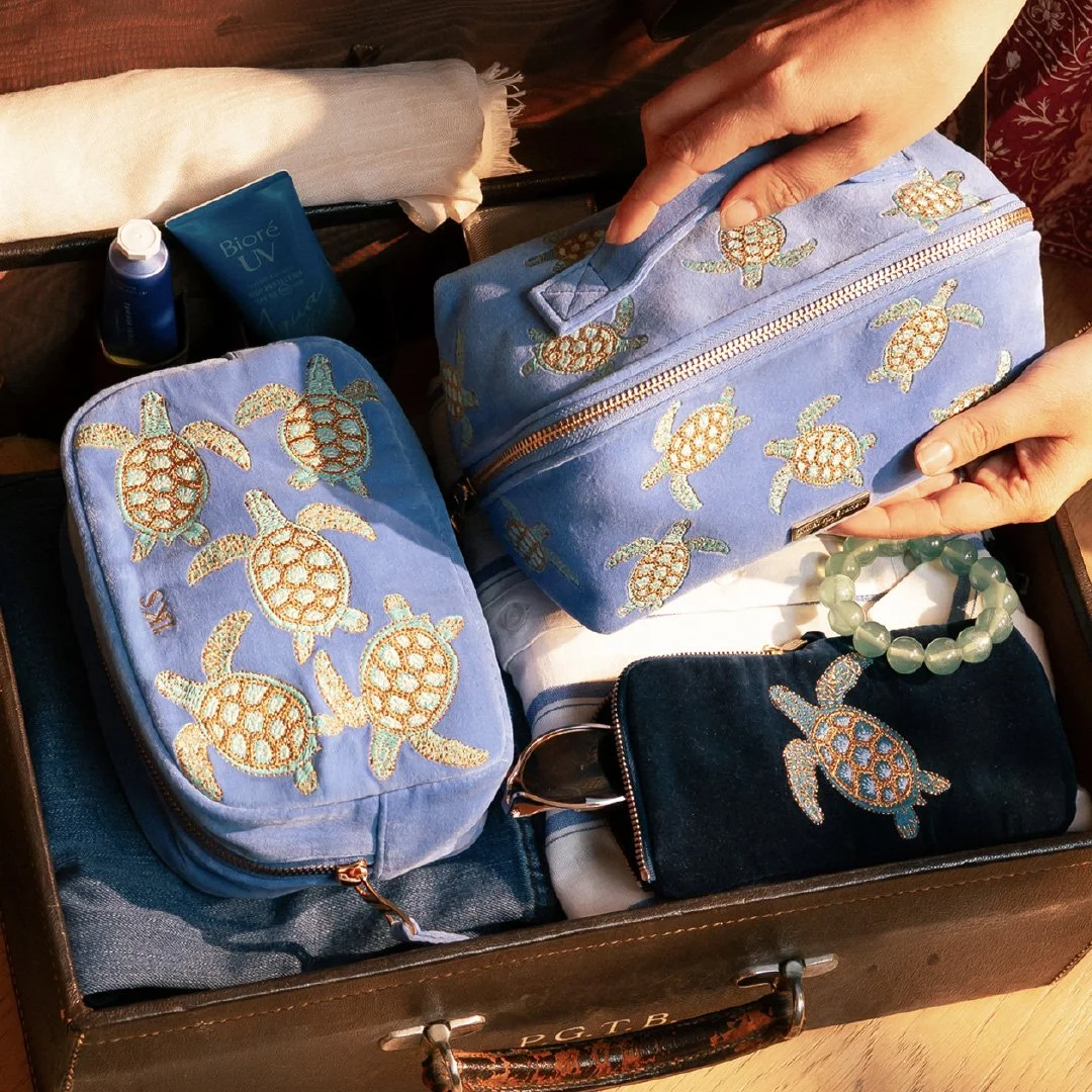 A suitcase with embroidered turtle designs contains travel essentials including two blue bags, a small black pouch, a green beaded bracelet, and toiletries.