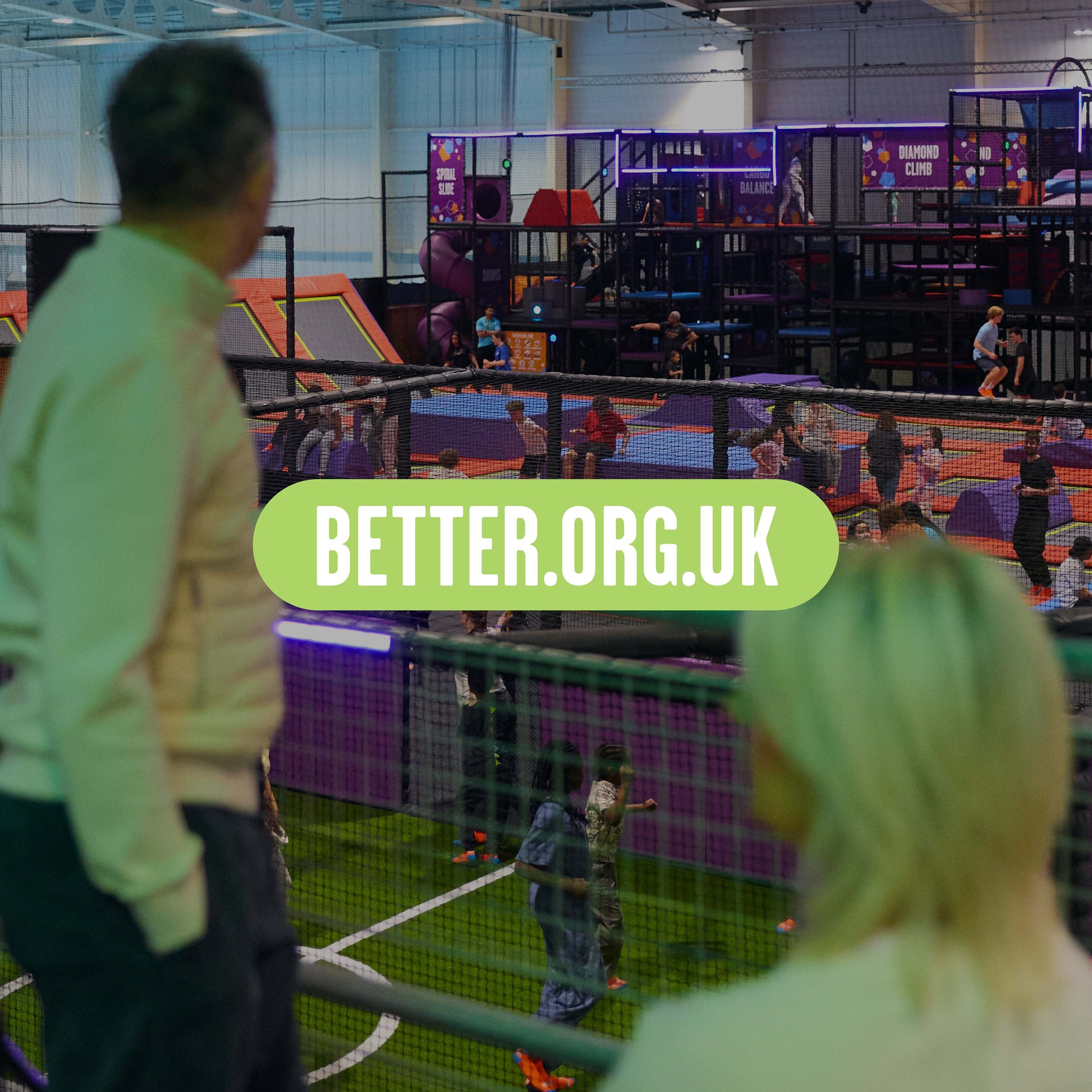 Indoor trampoline park with children jumping on trampolines, climbing structures, and slides, viewed through a safety net with a person in the foreground and industrial ceiling lighting.