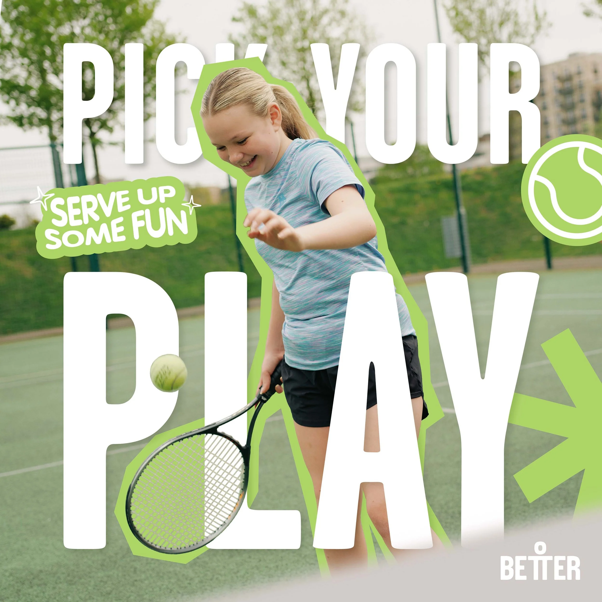 A young girl smiling while playing tennis outdoors on a sunny day, with green trees and buildings in the background.