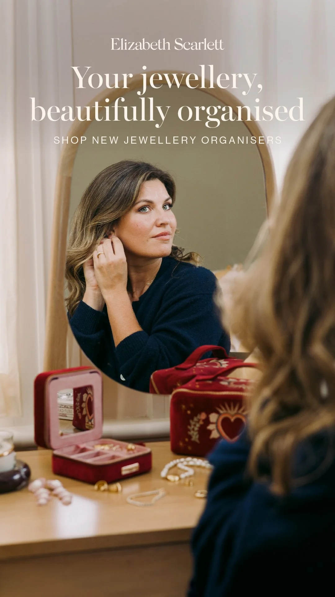 A woman looking in a mirror, with various jewellery items and storage cases on a table in front of her.