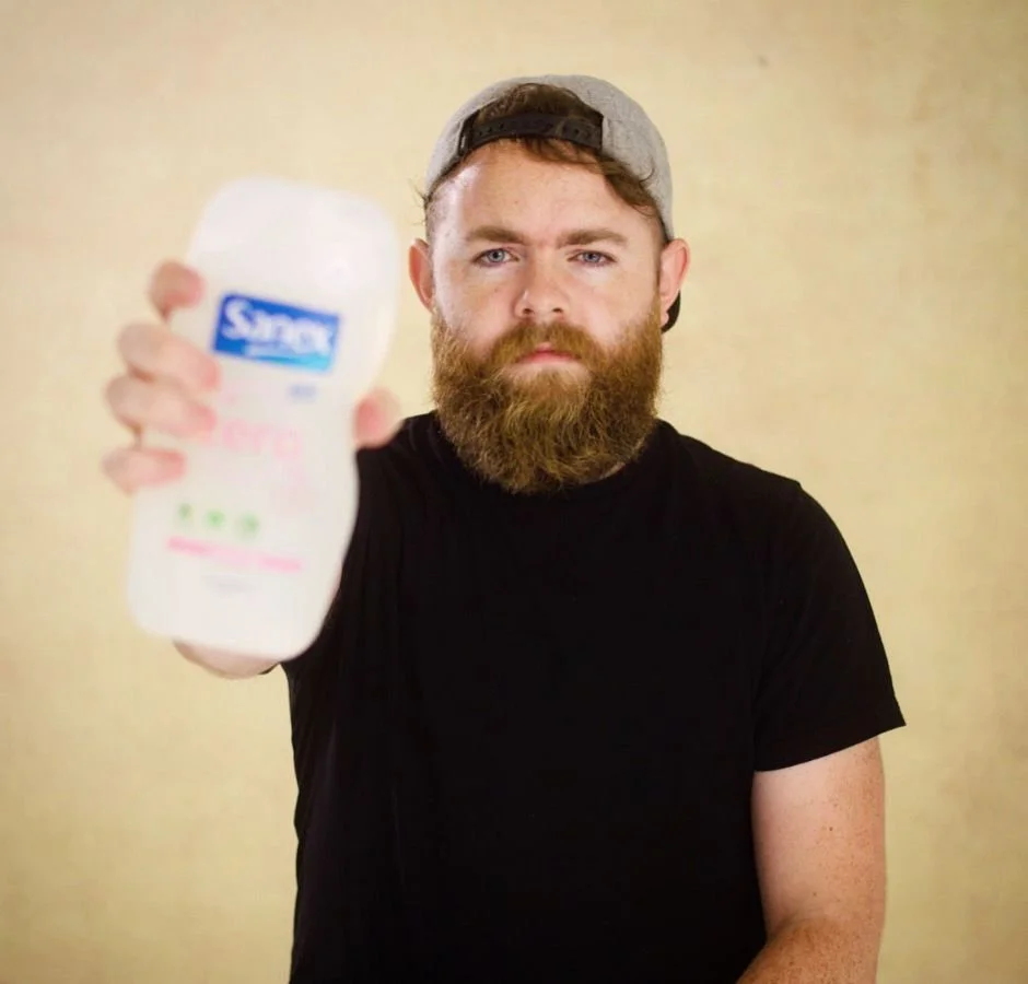 A bearded man wearing a gray cap backwards and a black T-shirt holds a bottle of Simple bodywash product towards the camera, with a neutral beige background.