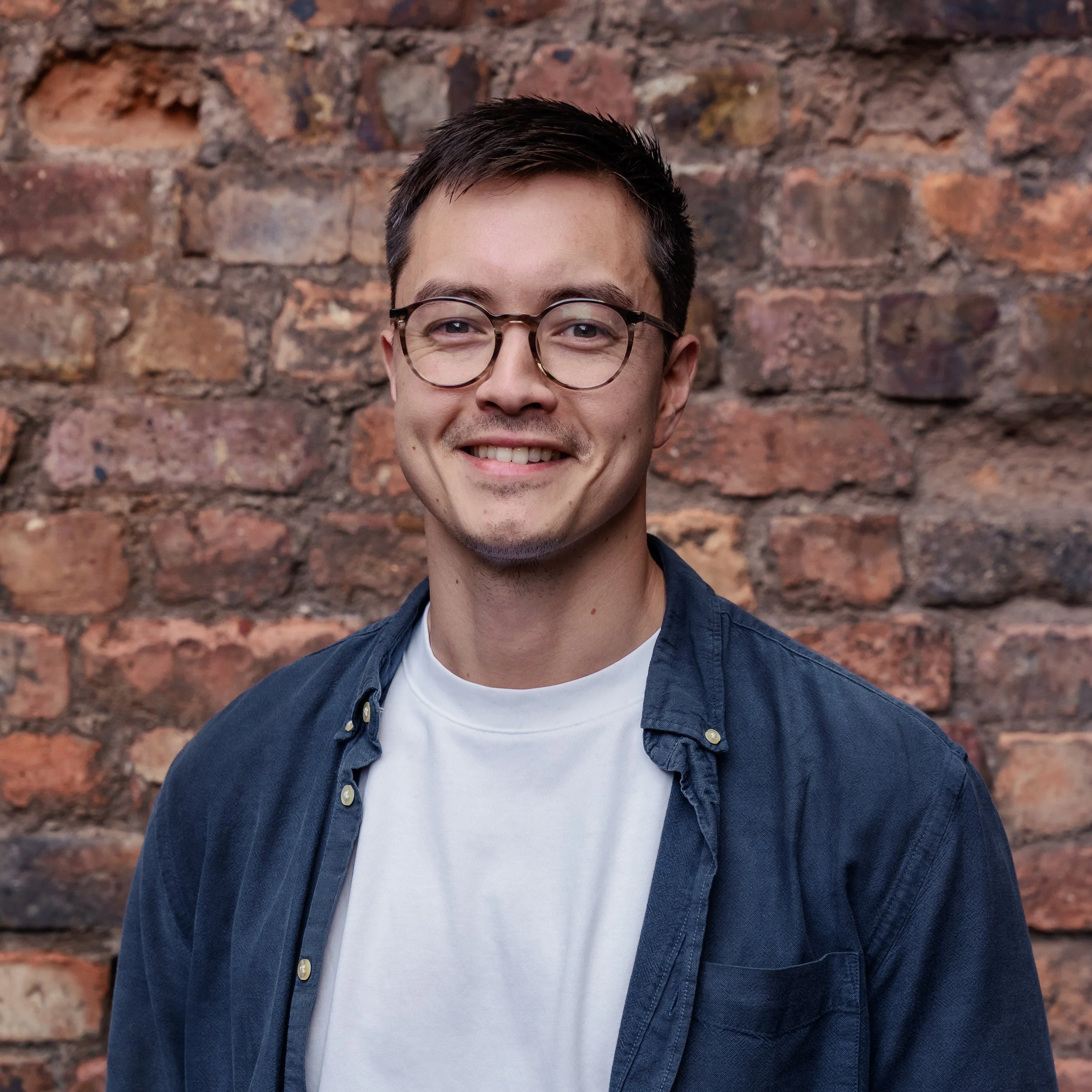 A young man with dark hair wearing glasses, a white t-shirt, and a blue shirt, smiling against a brick wall background.