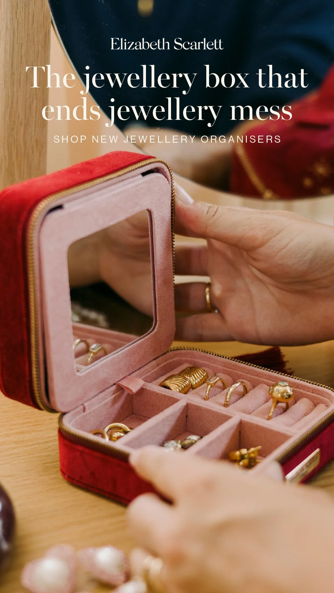 A hand holding open a pink velvet jewellery box with a mirror, containing various gold rings and earrings. In the background, a person wears a dark sweater