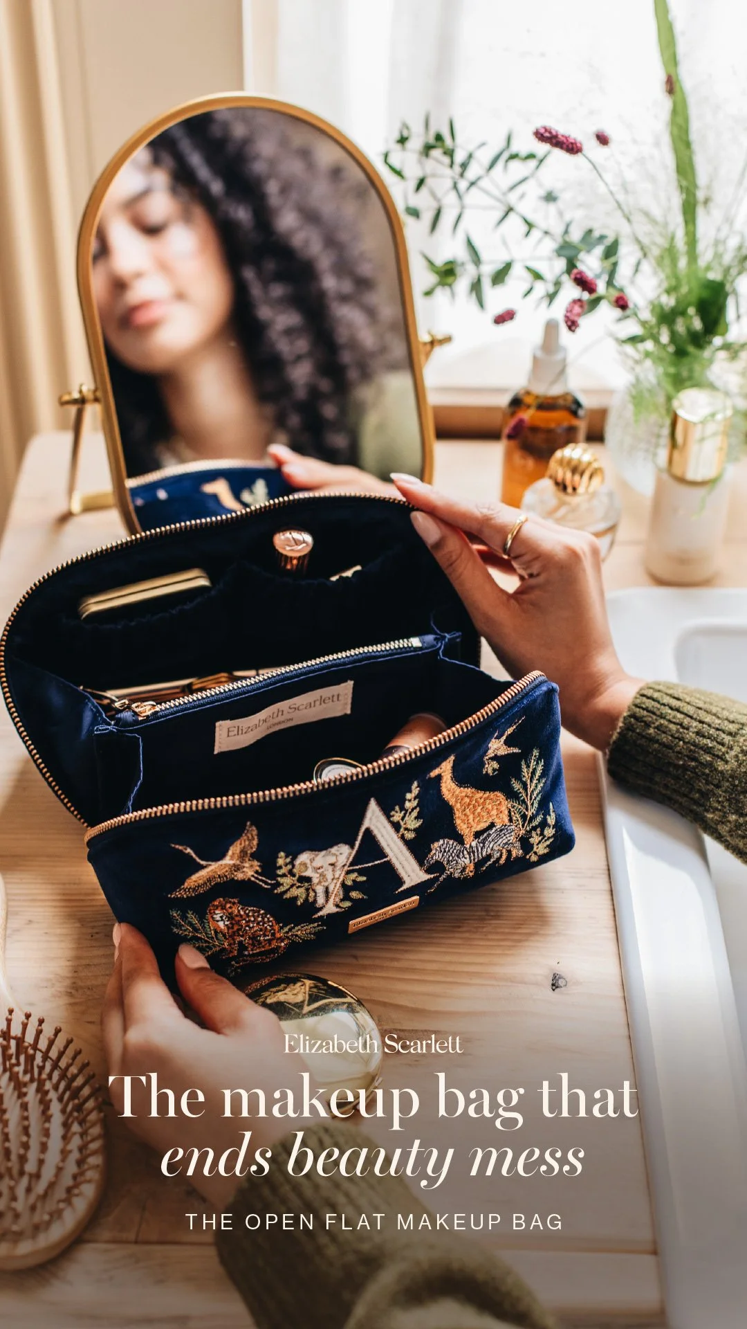 A person with curly hair looking into a mirror while reaching into a wildlife-themed makeup bag on a wooden table. The table has a small spray bottle, a pot of toothbrush, and decorative items, with flowers in the background.