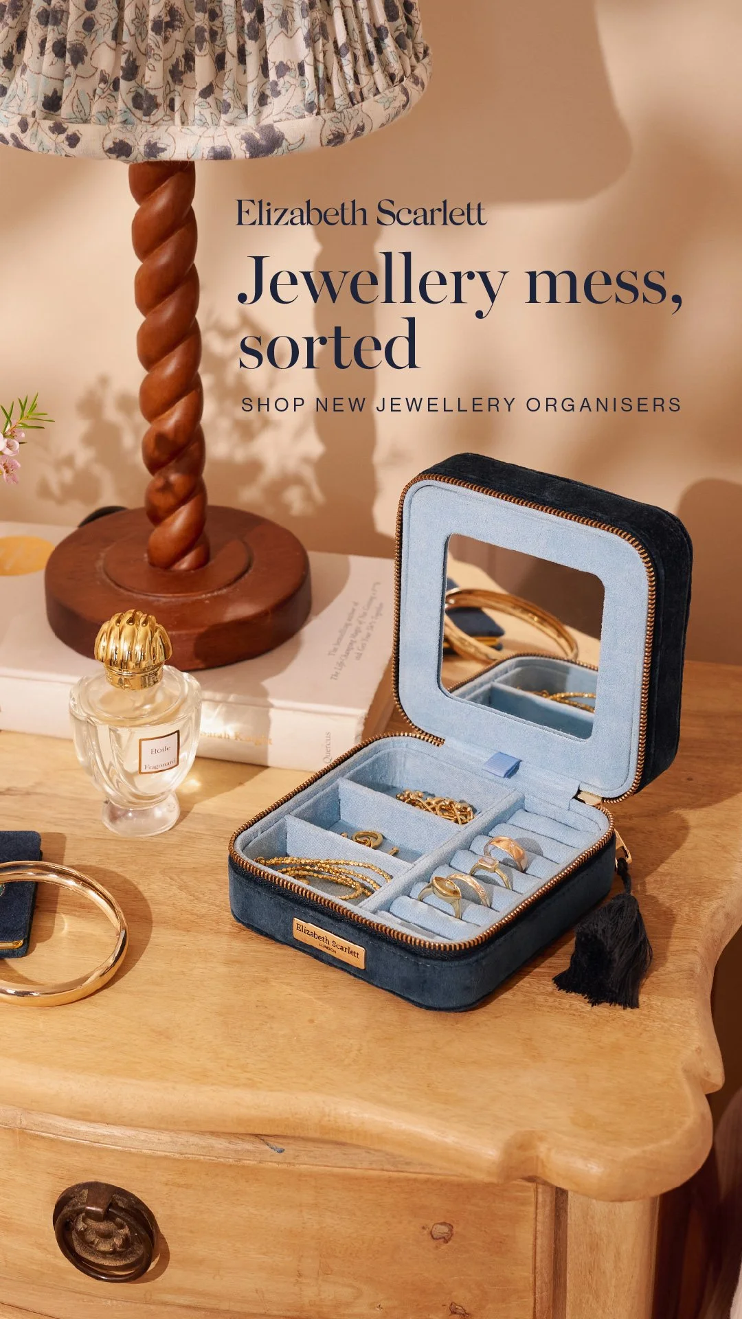 Open jewellery box with rings and necklaces, a perfume bottle, and other jewelry on a wooden surface, with a table lamp and books in the background.