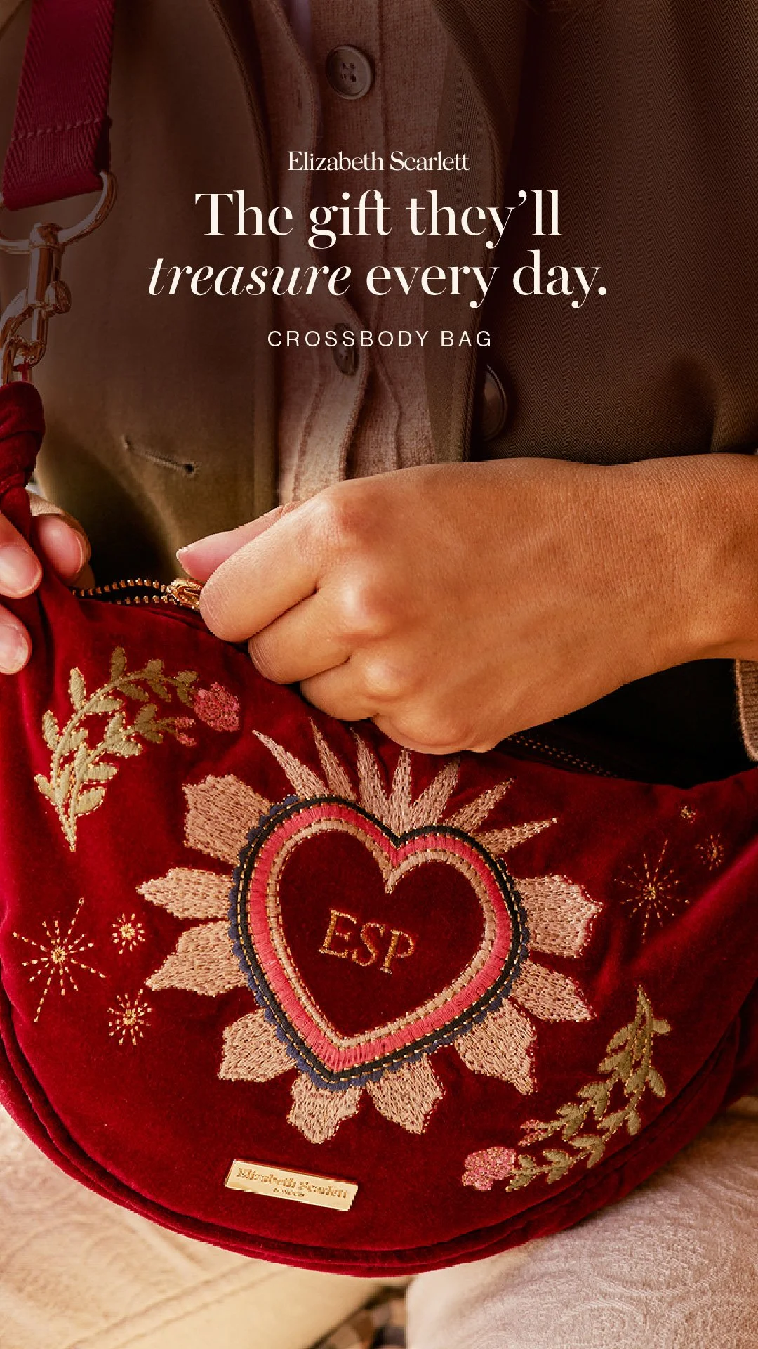 A person's hand holding a red embroidered crossbody bag with floral and heart designs, and initials ESP inside a heart, with a park or outdoor area in the background.