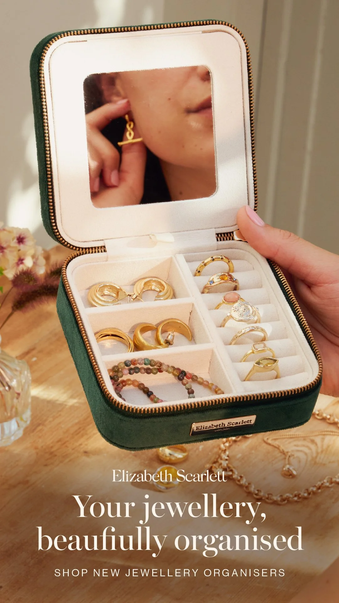A jewellery box with a mirror showing a woman’s face and hand. Inside are gold rings on display, with a beaded bracelet and other jewelry on a wooden surface.