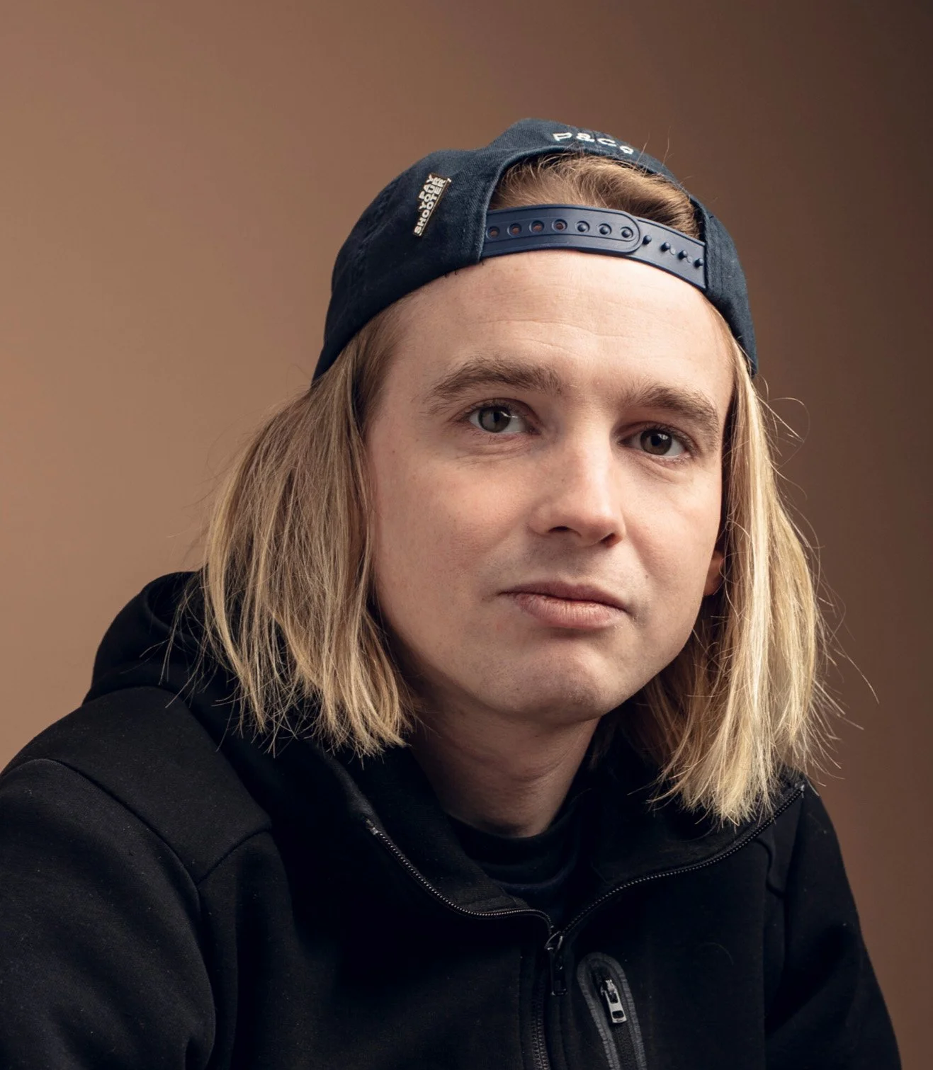 A young man with shoulder-length blond hair wearing a black baseball cap backward and a black zip-up jacket, looking at the camera against a brown background.