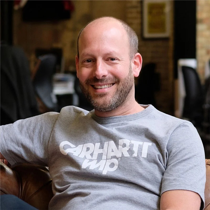 A smiling man with a beard and bald head, wearing a gray Carhartt Work in Progress t-shirt, sitting on a leather couch in an office setting.