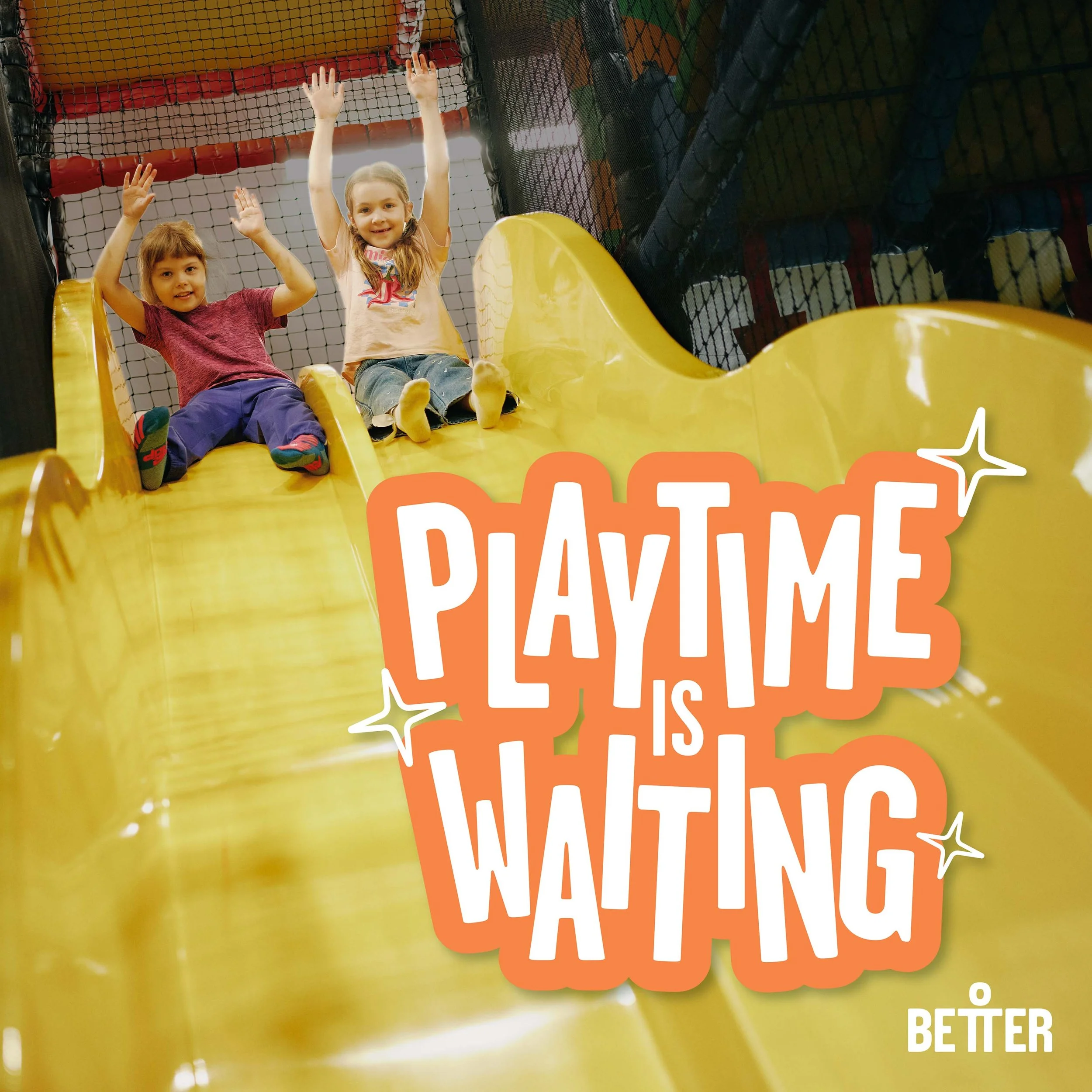 Two young girls are sitting at the top of a yellow playground slide, smiling and waving. Behind them is black safety netting and a structure with colorful play equipment.
