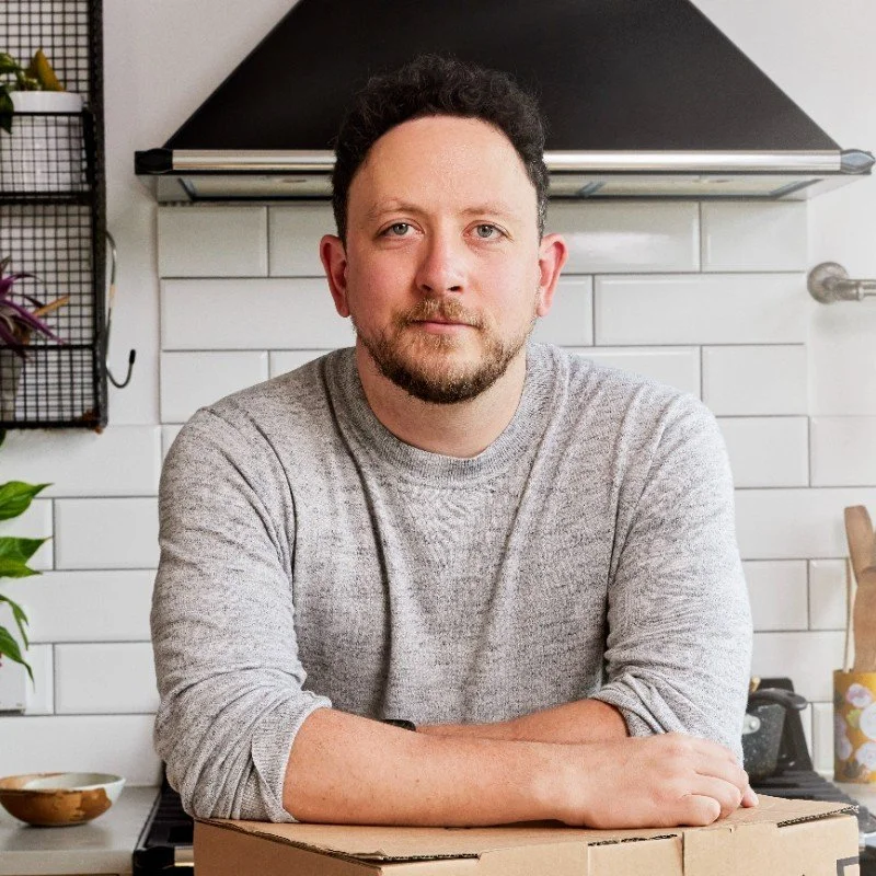 Man with curly hair and beard sitting at a kitchen counter with a cardboard box, white tiled backsplash, and kitchen appliances in the background.