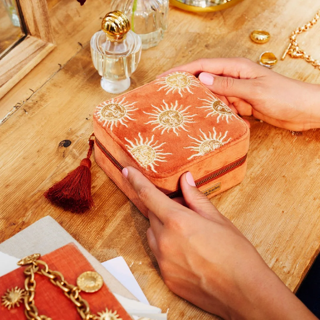 A person's hands are holding an orange velvet jewellery box with sun designs on top. The box is on a wooden table with perfume bottles, jewelry, and a mirror nearby.