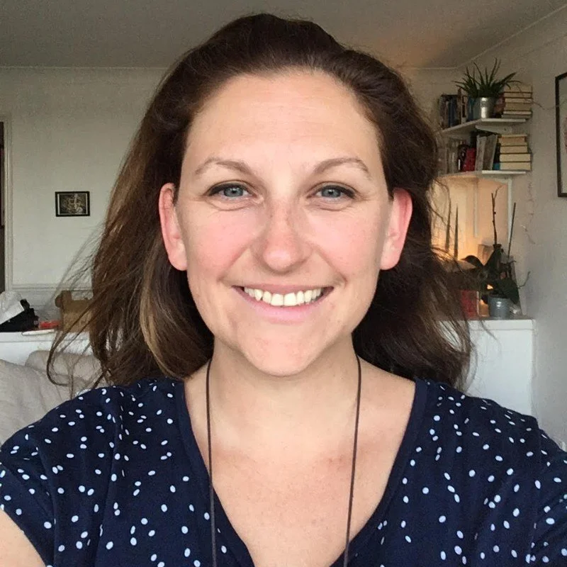 A woman with brown hair and blue eyes smiling in a room with books and plants in the background.