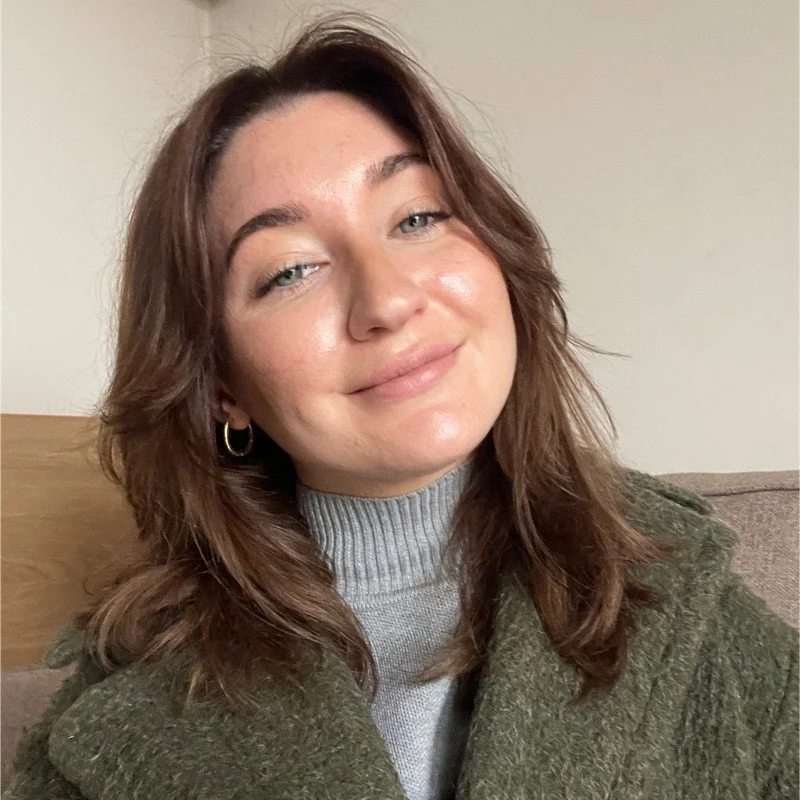 A young woman with brown hair and blue eyes smiling, wearing a gray turtleneck and an olive green sweater, sitting on a beige couch indoors.