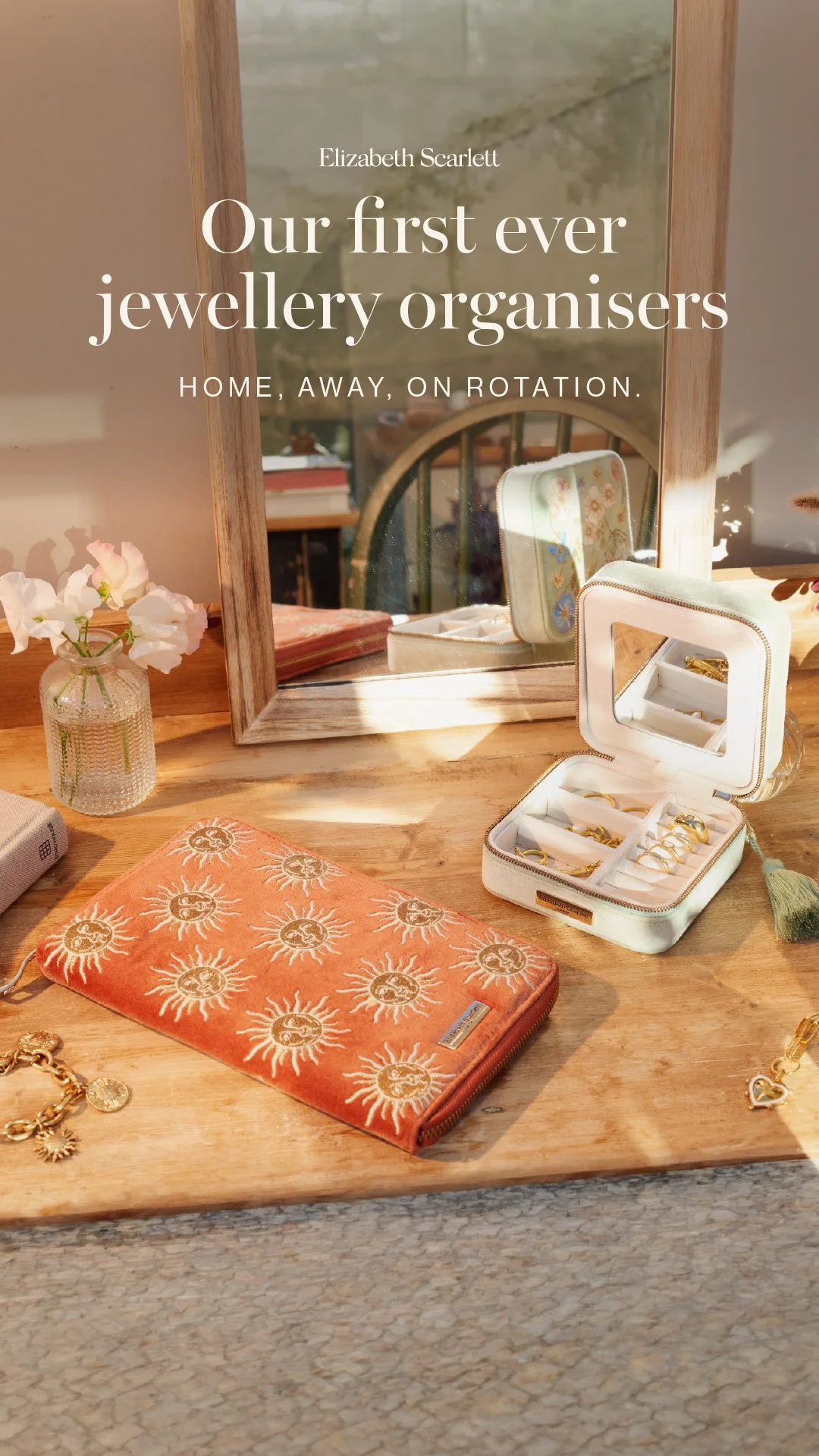 A wooden dressing table with a large mirror, a small vase with pink flowers, a closed orange jewellery case with a sun design, and an open white jewellery box containing rings and earrings, with sunlight shining on the table.