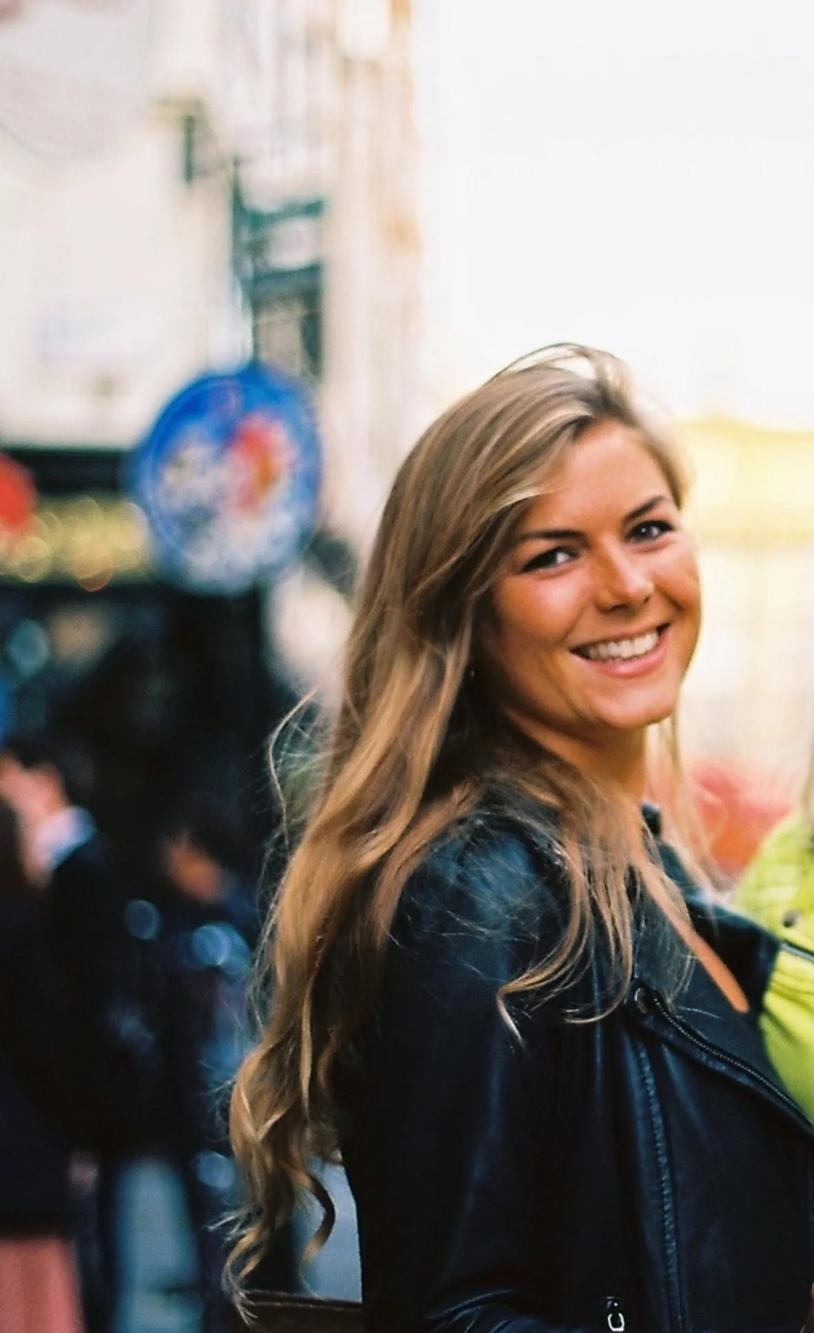 A young woman with long wavy hair smiling in a crowded outdoor setting.