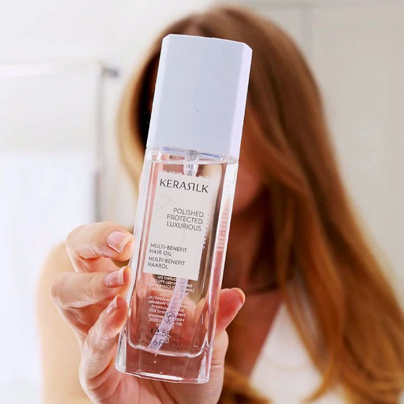 Woman with strawberry blonde hair holding a bottle of Kerasilk hair oil in front of her face.