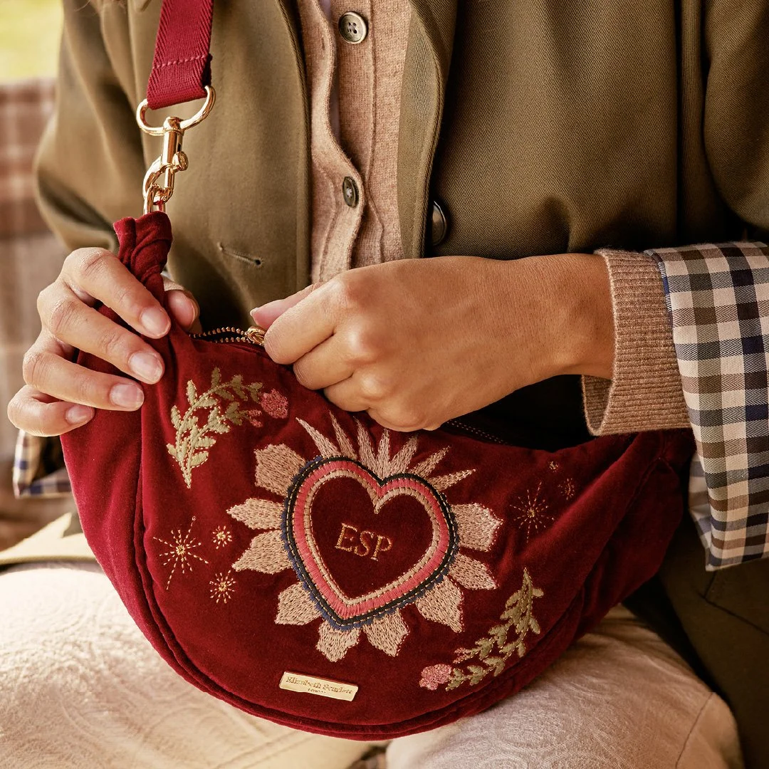 Person holding a red velvet crossbody bag with embroidered heart, sunburst, and floral design, featuring the initials 'ESP' and a gold label.