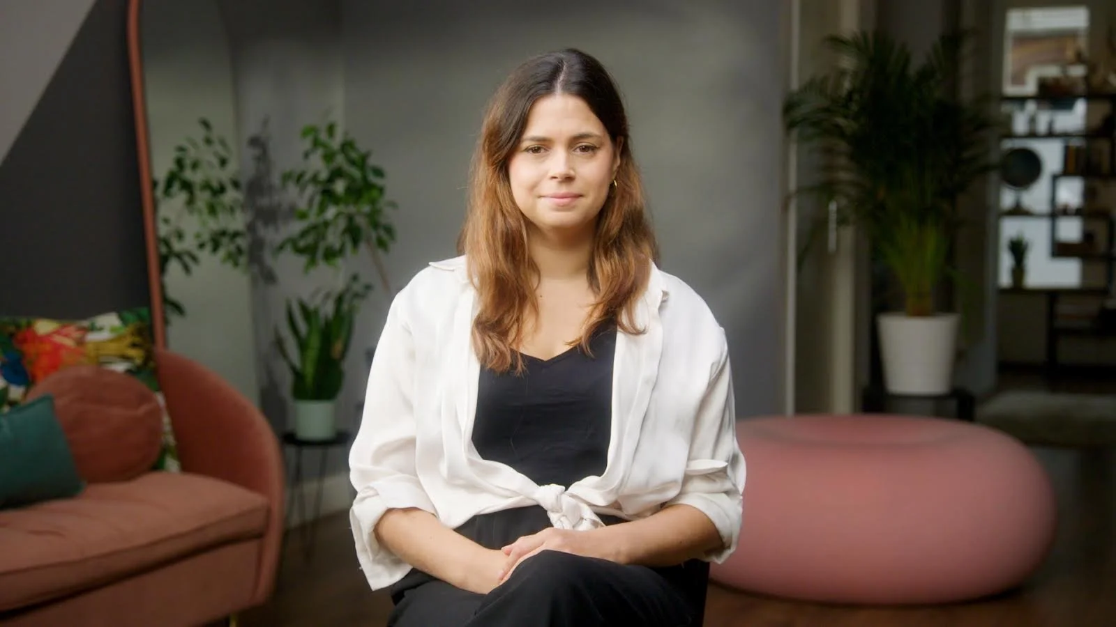 A woman with shoulder-length brown hair sitting on a chair in a cozy living room with plants and modern decor.