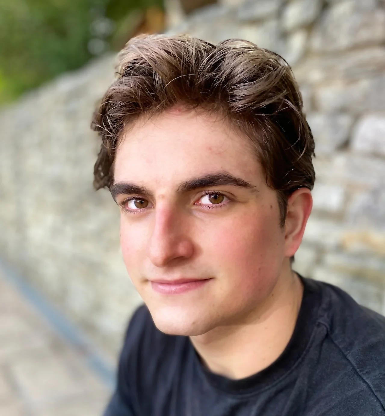 Close-up of a young man with brown hair and hazel eyes, smiling gently, outdoors against a stone wall background