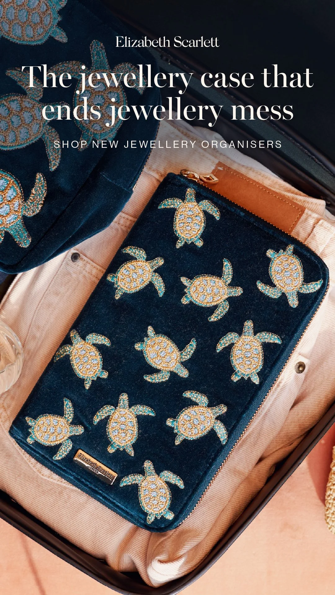 A navy blue velvet jewellery case with turtle embroidery, placed in an open suitcase with light-colored clothing inside.