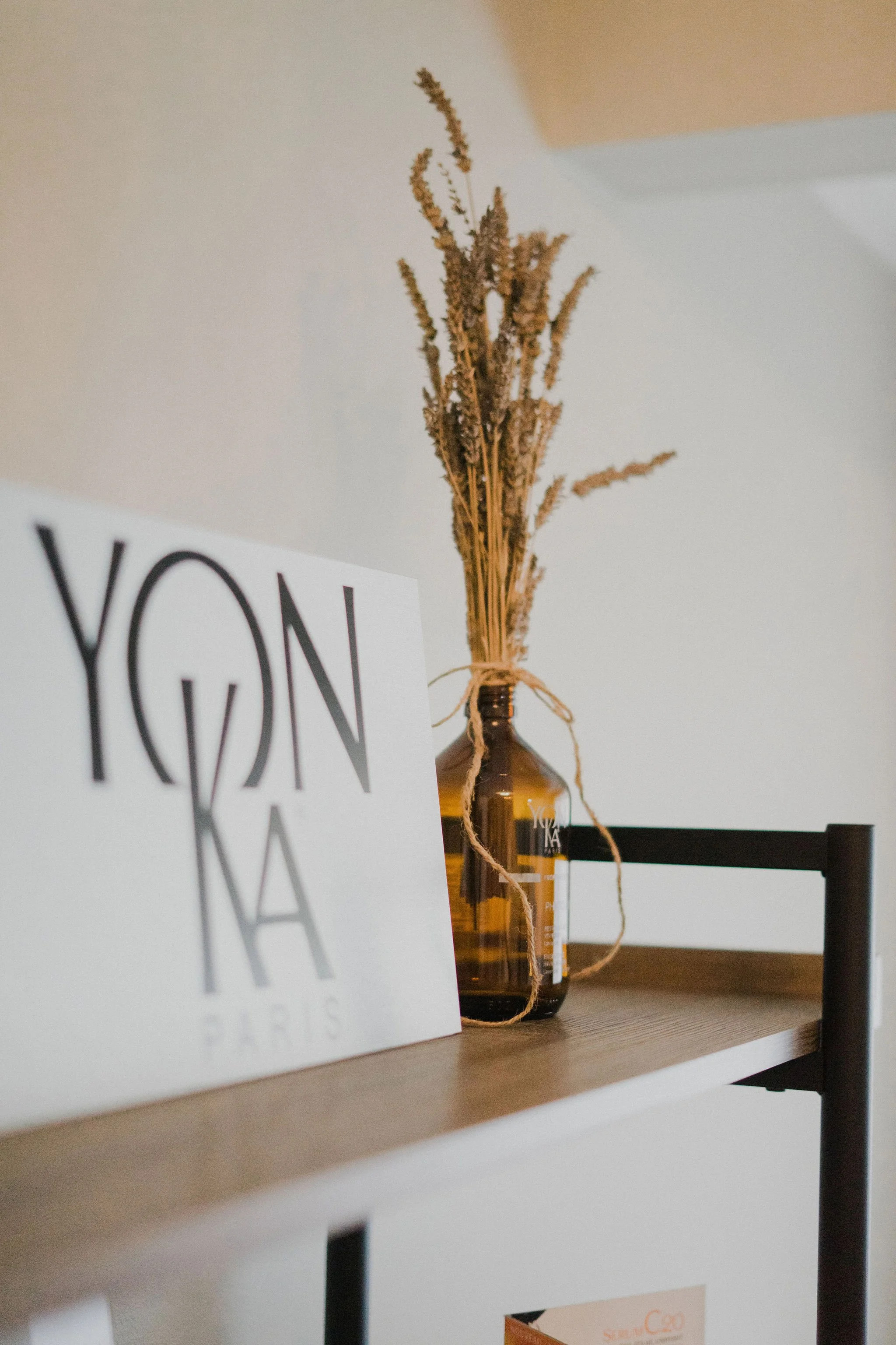 A tabletop with a brown glass bottle holding dried lavender, a white card with black text partially visible that reads 'YON', and a black stand supporting a white paper with orange text.