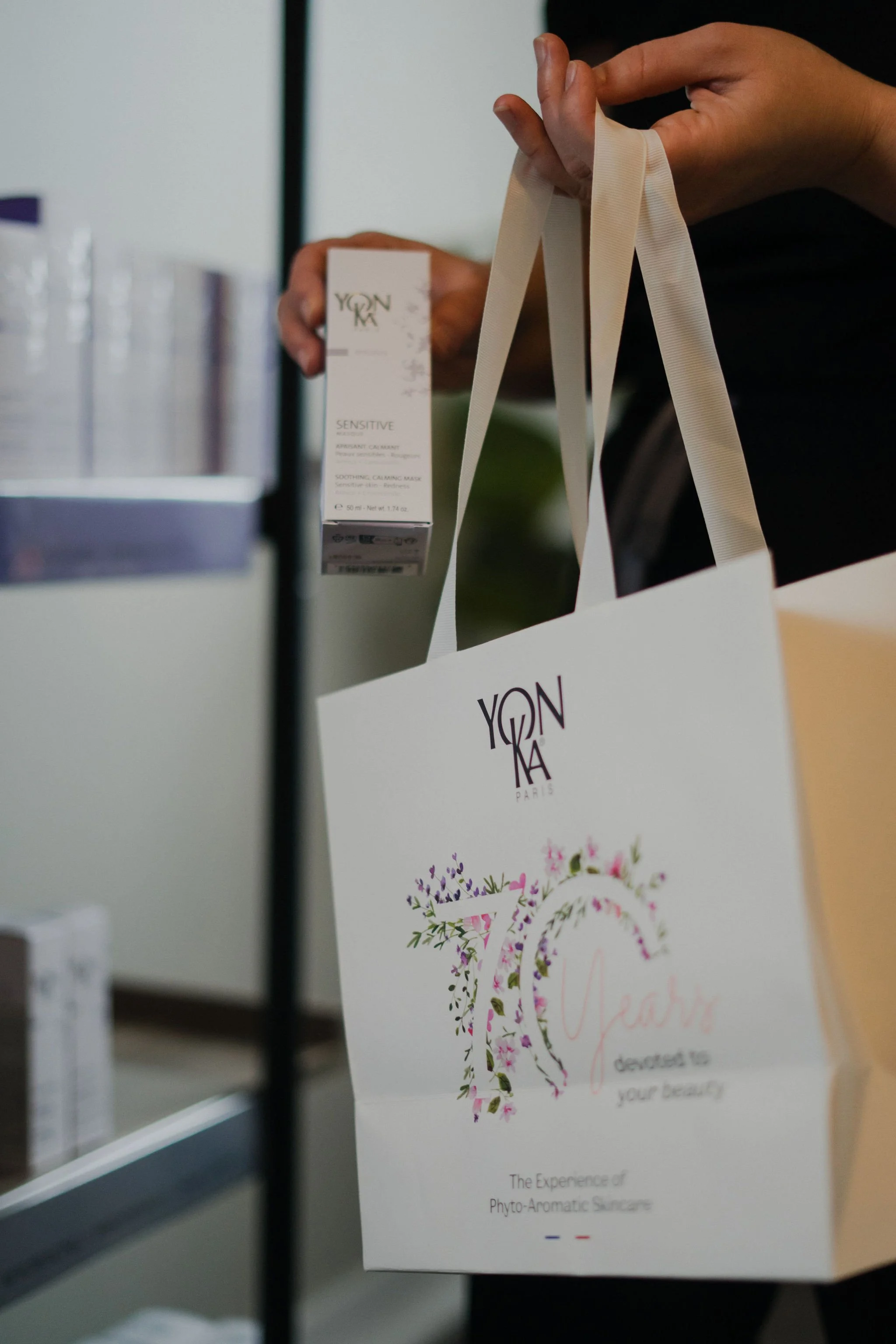 A person holding a white box labeled 'YONKA' and the word 'Sensitive'. The box appears to be for a calming and soothing skincare mask. The person is also holding a white shopping bag with the same 'YONKA' logo and decorative floral text that says 'Bi