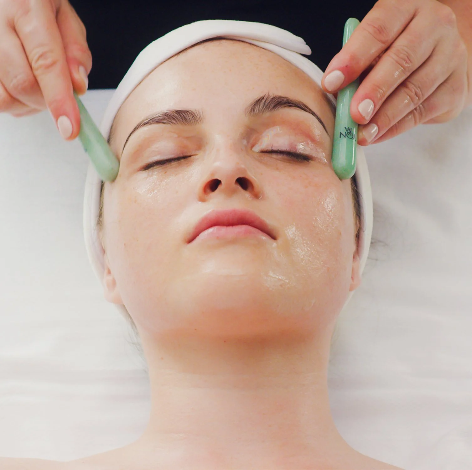 Woman receiving a facial treatment with jade rollers on her face, lying down with eyes closed.