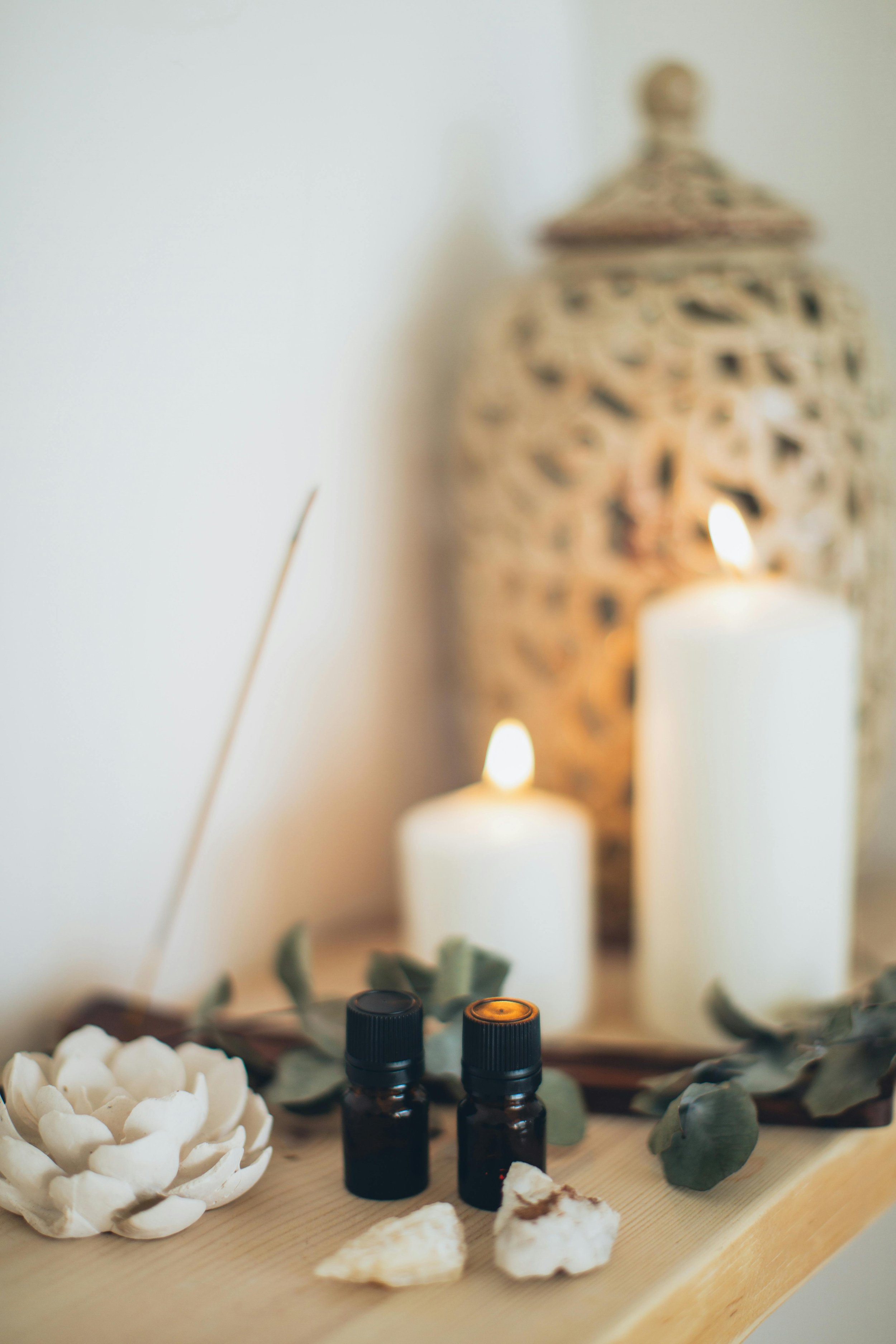 A close-up of two small black essential oil bottles with orange and black caps on a wooden surface. Behind them are white candles and a decorative ceramic pine cone. A large ornate jar with a lid is in the background, along with green eucalyptus leav