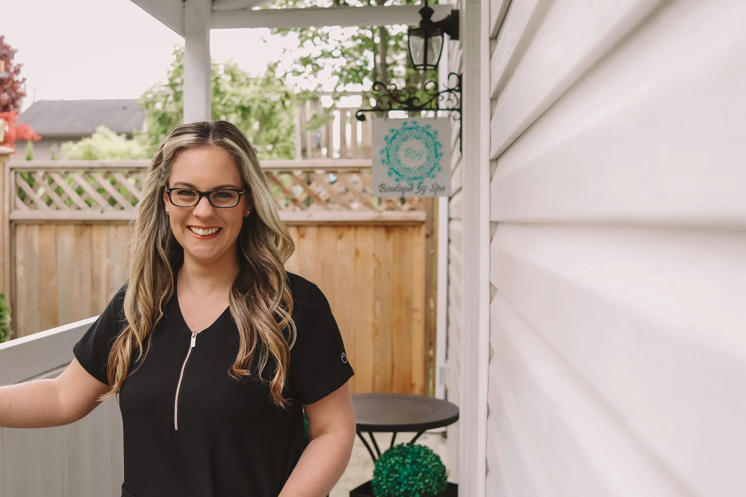 A woman with long, wavy hair and glasses smiling, standing outside near a white house with a wooden fence and a sign that reads 'Beauty & Spa.'