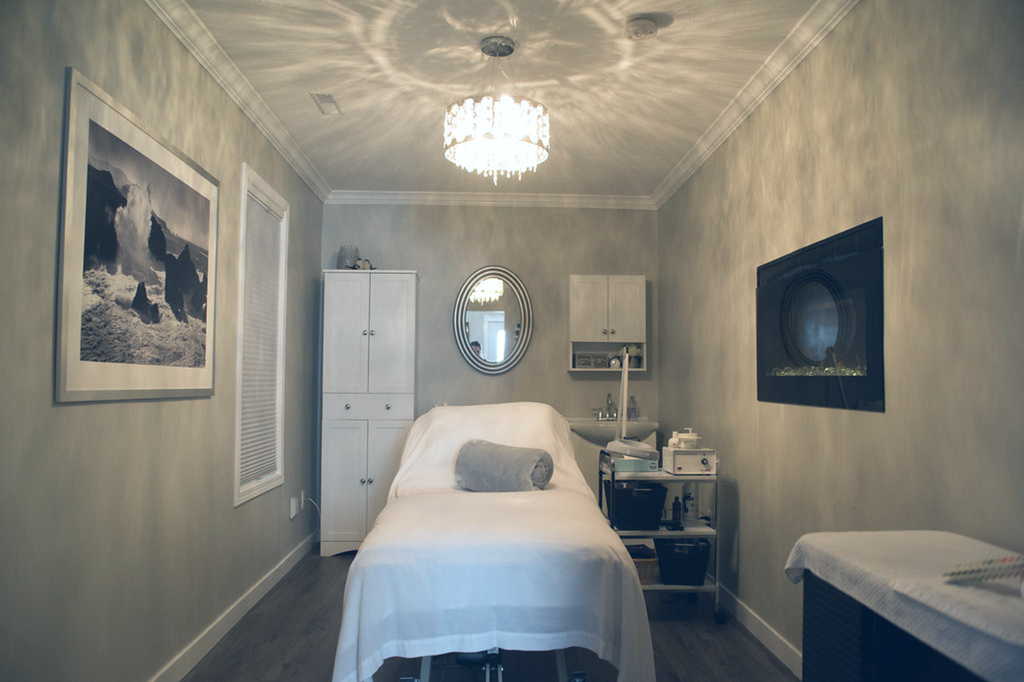 A room with a massage bed, a white cabinet, a wall-mounted mirror, a small white storage cart, a wall-mounted television, and framed artwork, illuminated by a chandelier.