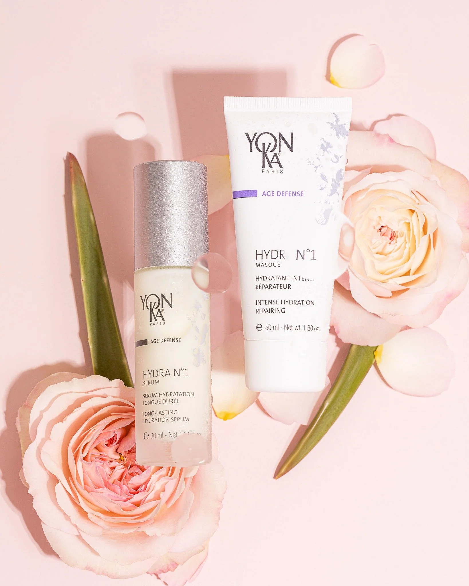 Two skincare products from Yonka Paris, a serum and a hydrating mask, placed on a pink background with pink roses and green leaves.