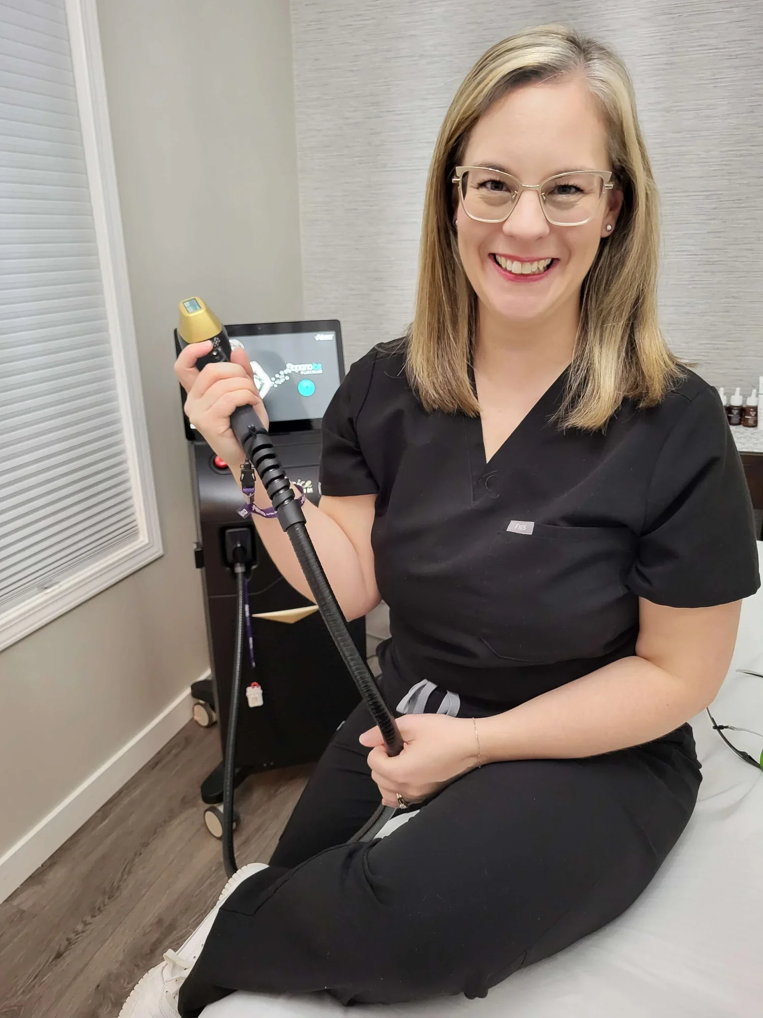 A smiling woman in a black medical uniform holding a handheld laser hair removal device in a clinical setting.