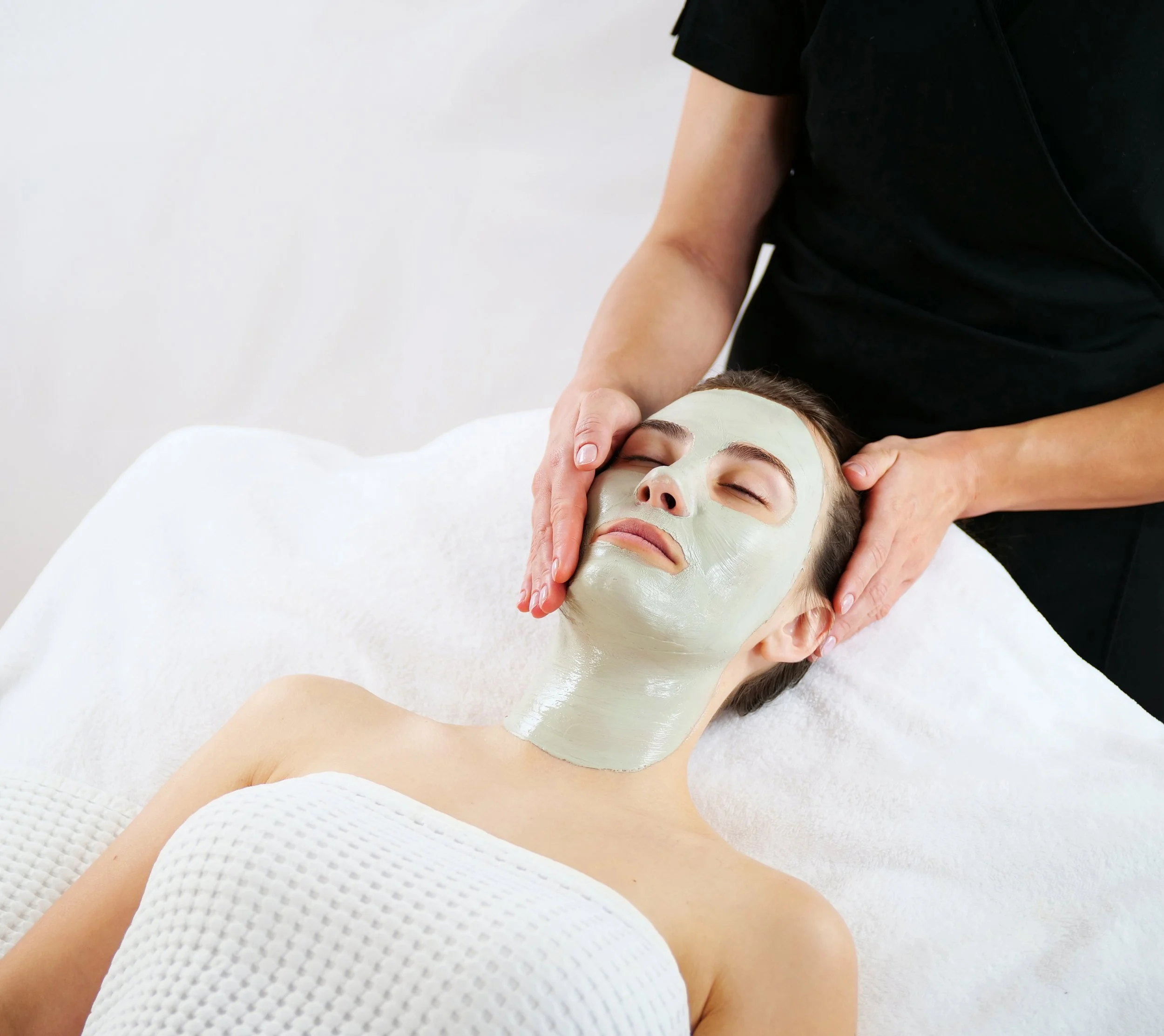 A woman receiving a facial treatment with a green facial mask applied, lying down with eyes closed, at a spa or salon.
