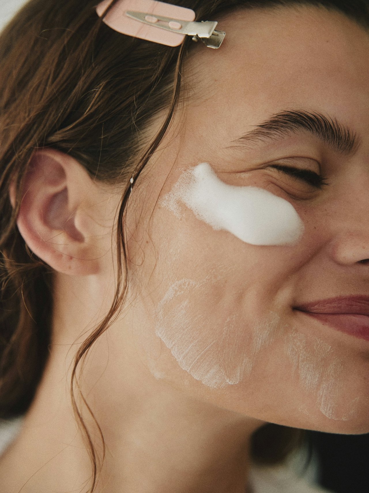 Woman with foam on cheek, smiling, with hair clipped back.