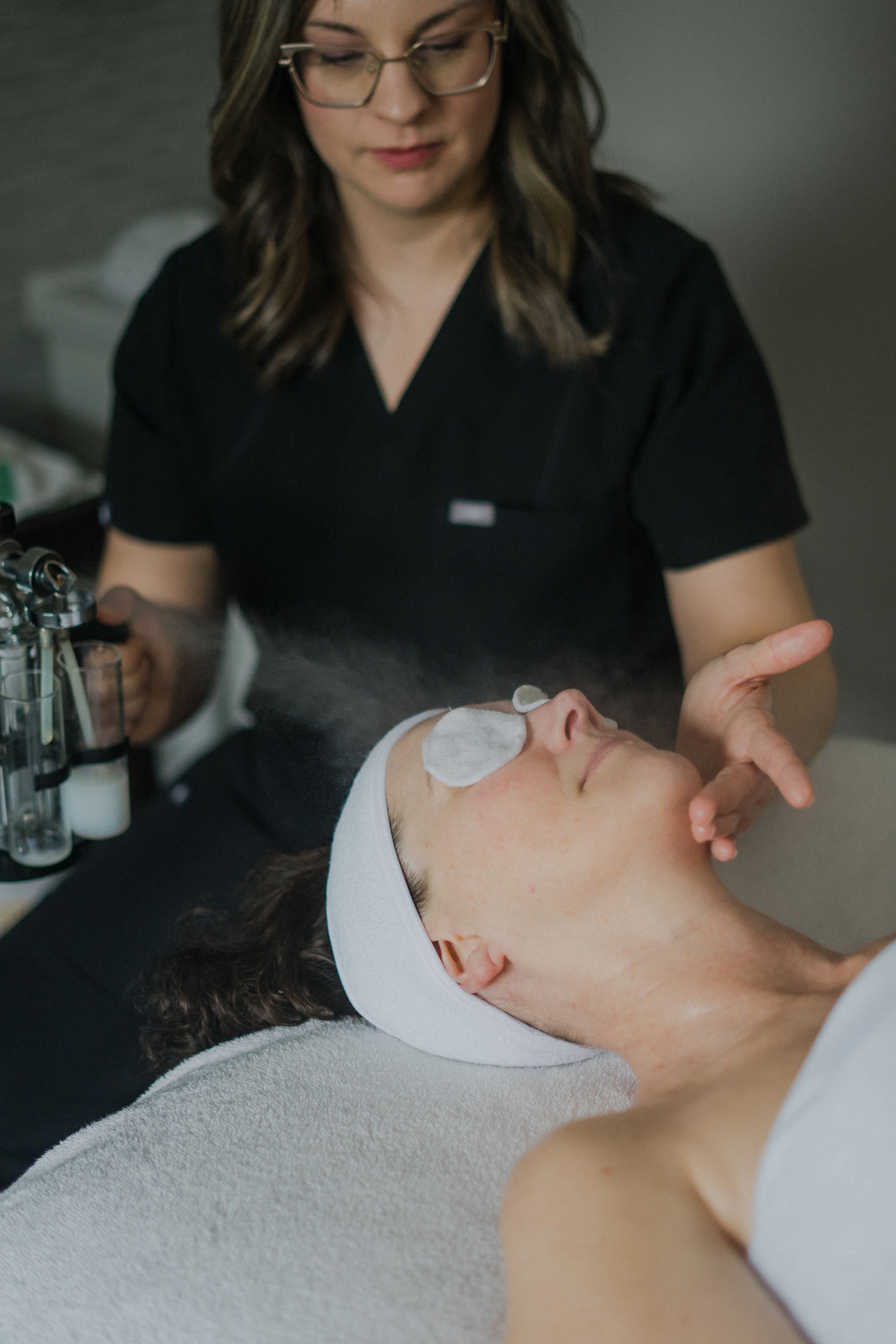 A woman in a black uniform, wearing glasses, is performing a facial treatment on a woman lying down with her eyes closed and a towel covering her chest. The woman receiving the treatment has eye pads on her eyes and is relaxed.
