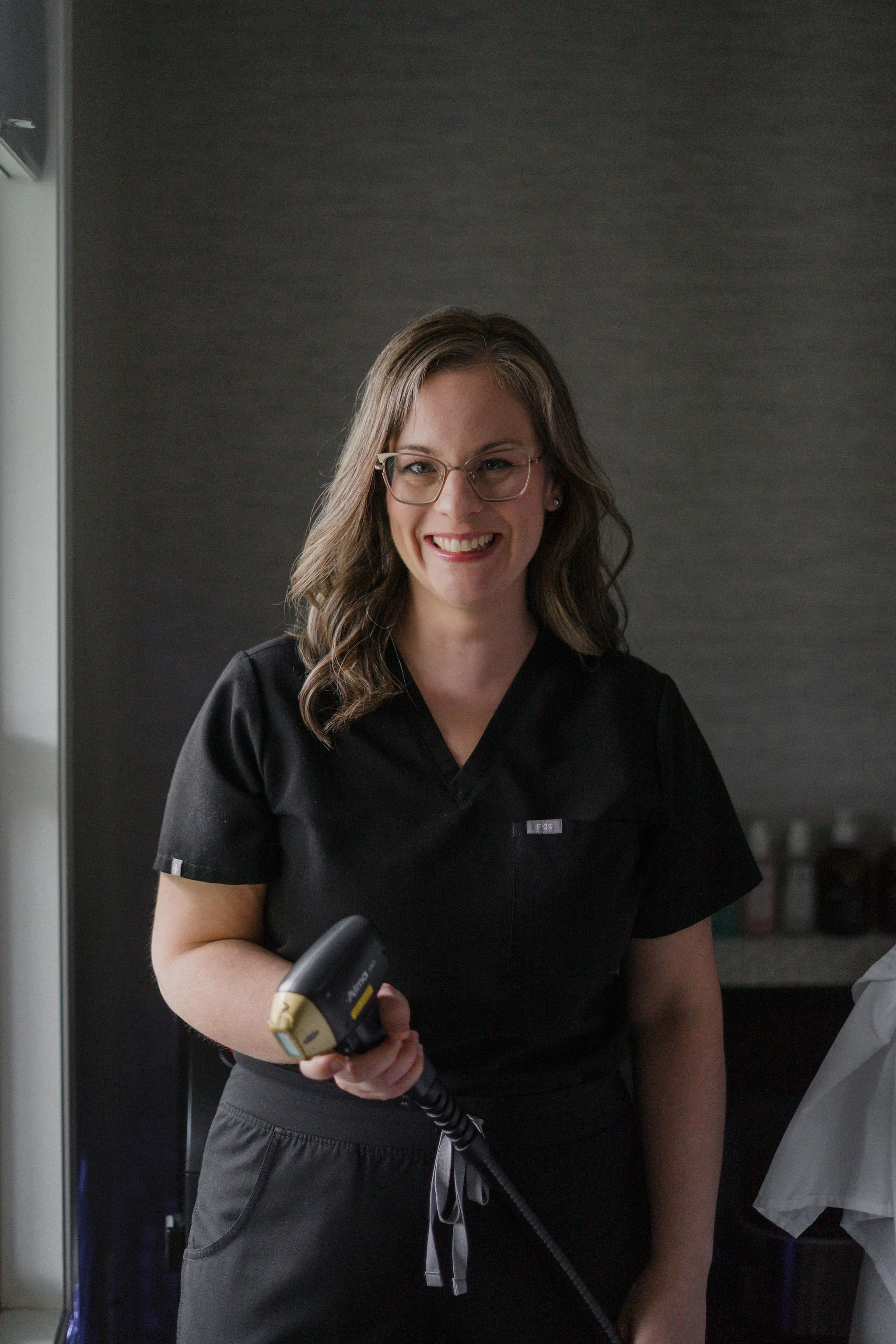 A woman in black scrubs smiling while holding a laser hair remover, standing in a professional environment.