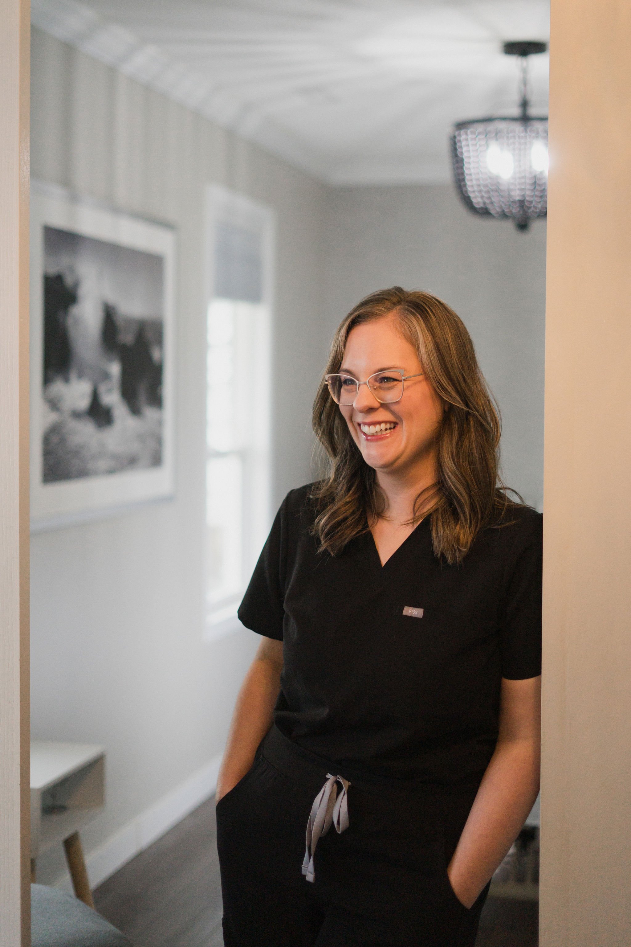 A woman with long brown hair and glasses smiling, wearing a black shirt with a small nametag, standing in a room with a chandelier and black and white artwork on the wall.
