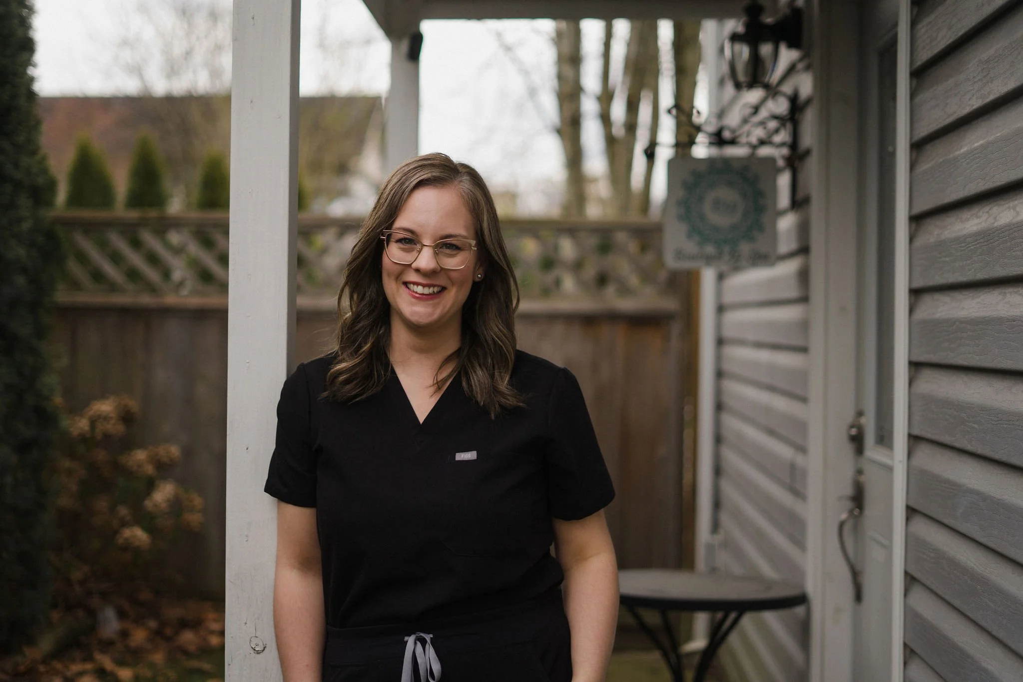 A young woman with long brown hair, glasses, and a black shirt smiling outdoors near a house with gray siding, trees, and a white fence in the background.