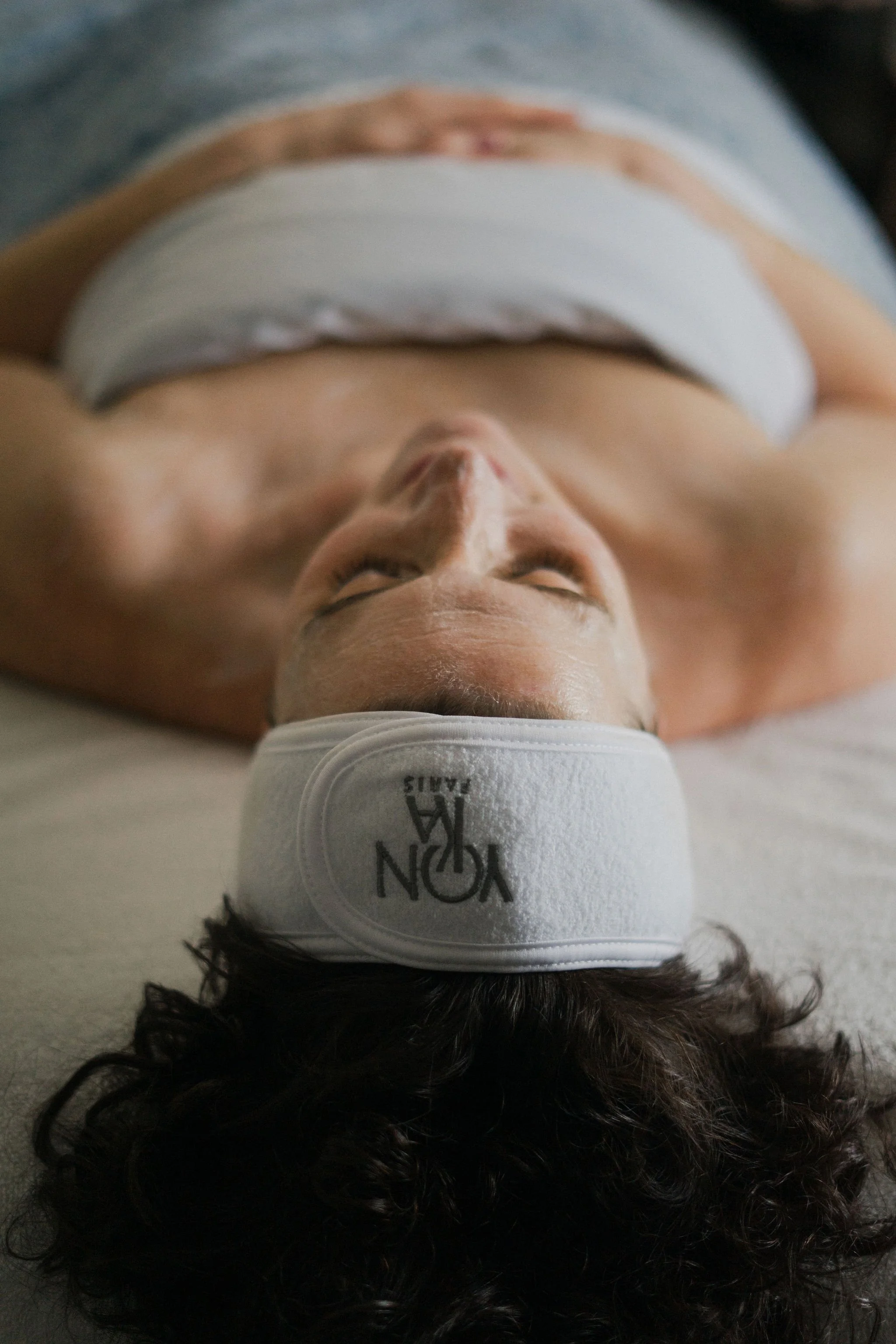 A person with dark, curly hair lying on their back, wearing a white headband with black text and a white tank top.