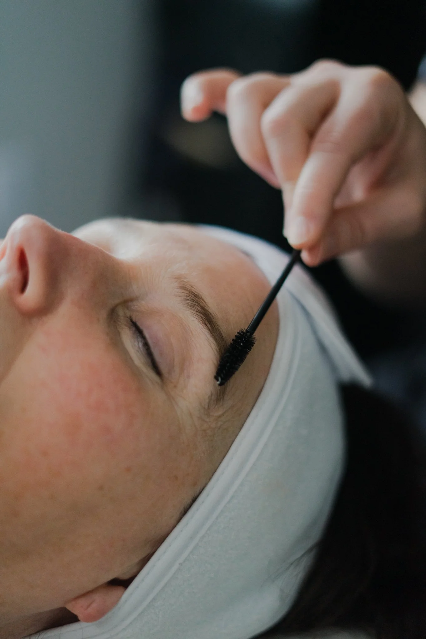 A person lying down with eyes closed, getting a makeup brush applied to their forehead by another person, possibly during makeup application.