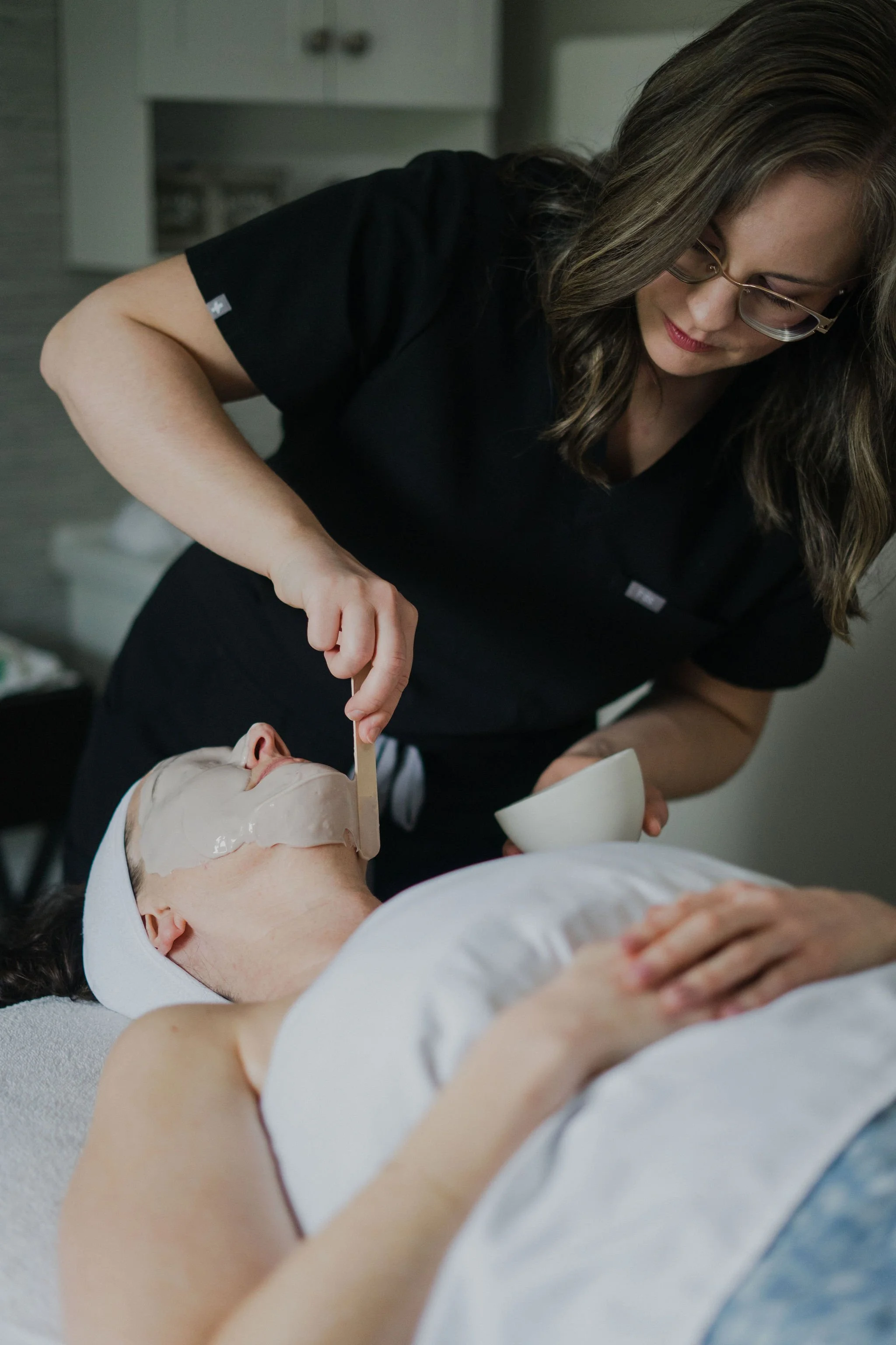 A woman is applying a facial mask to a person lying down with a towel on their head, in a skincare or beauty treatment setting.